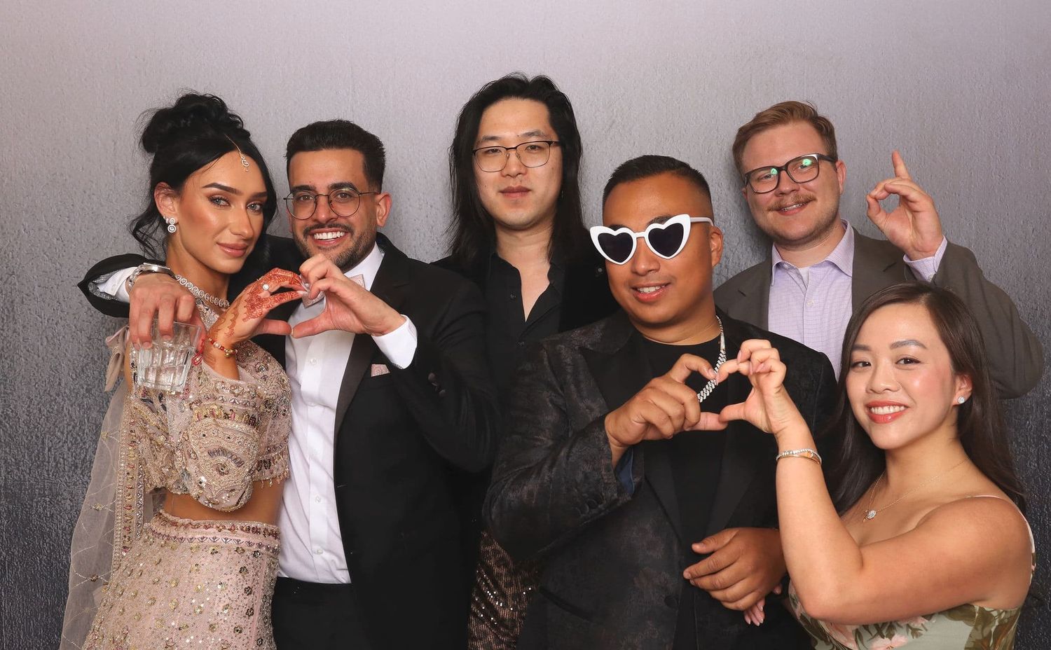 Group of six friends making heart shapes with hands at a photo booth event.
