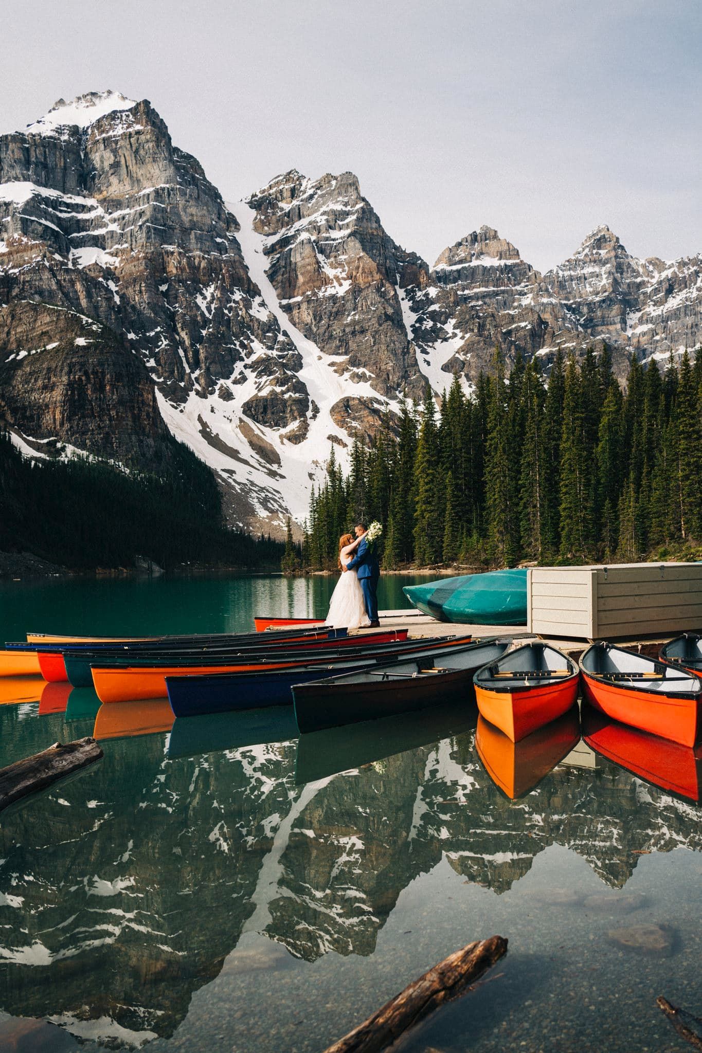 Couple embracing on a dock at Moraine Lake with colorful canoes, turquoise water, and snowy Rocky Mountains backdrop.