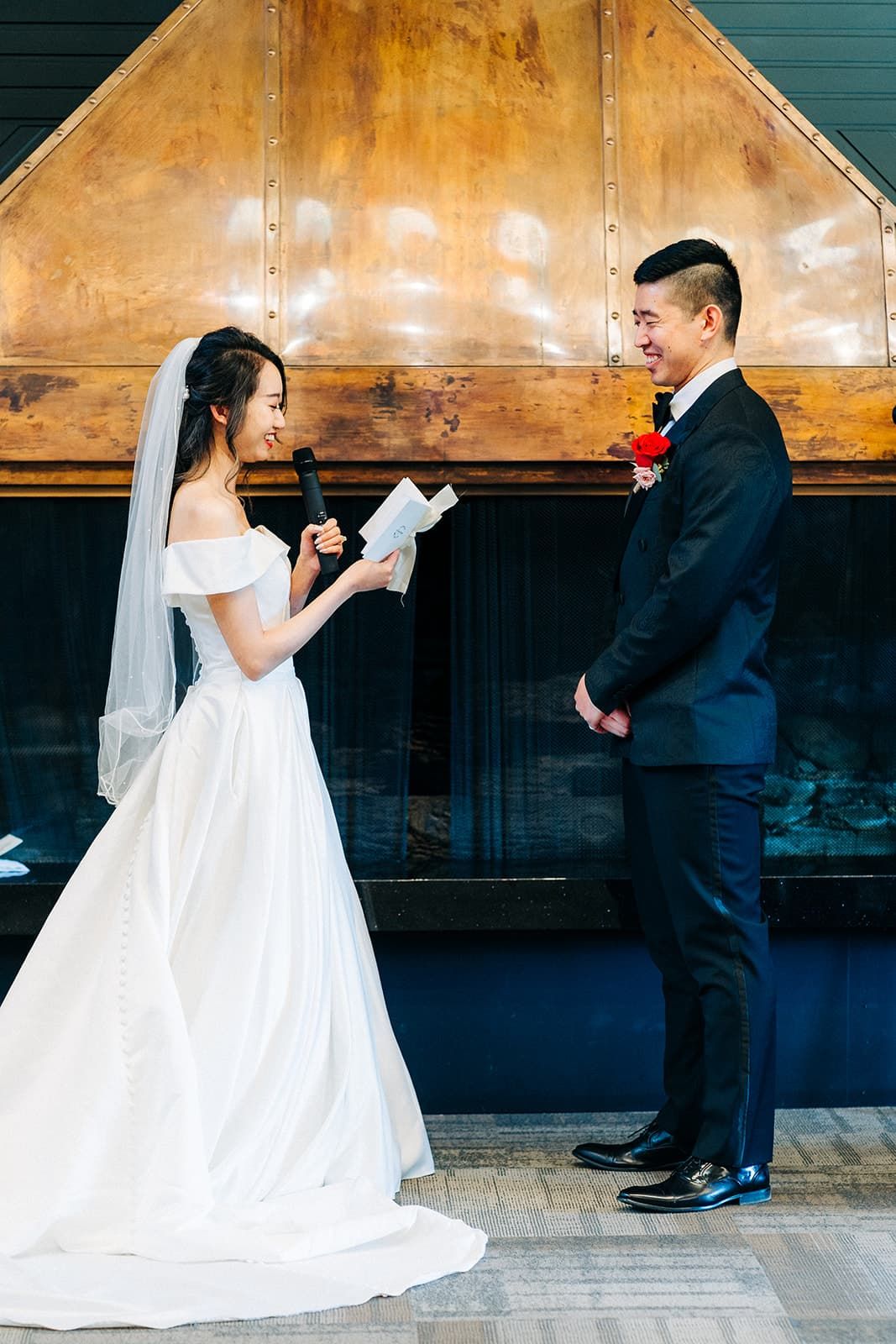 Bride reads wedding vows to smiling groom during intimate ceremony, both dressed elegantly in white gown and dark suit.