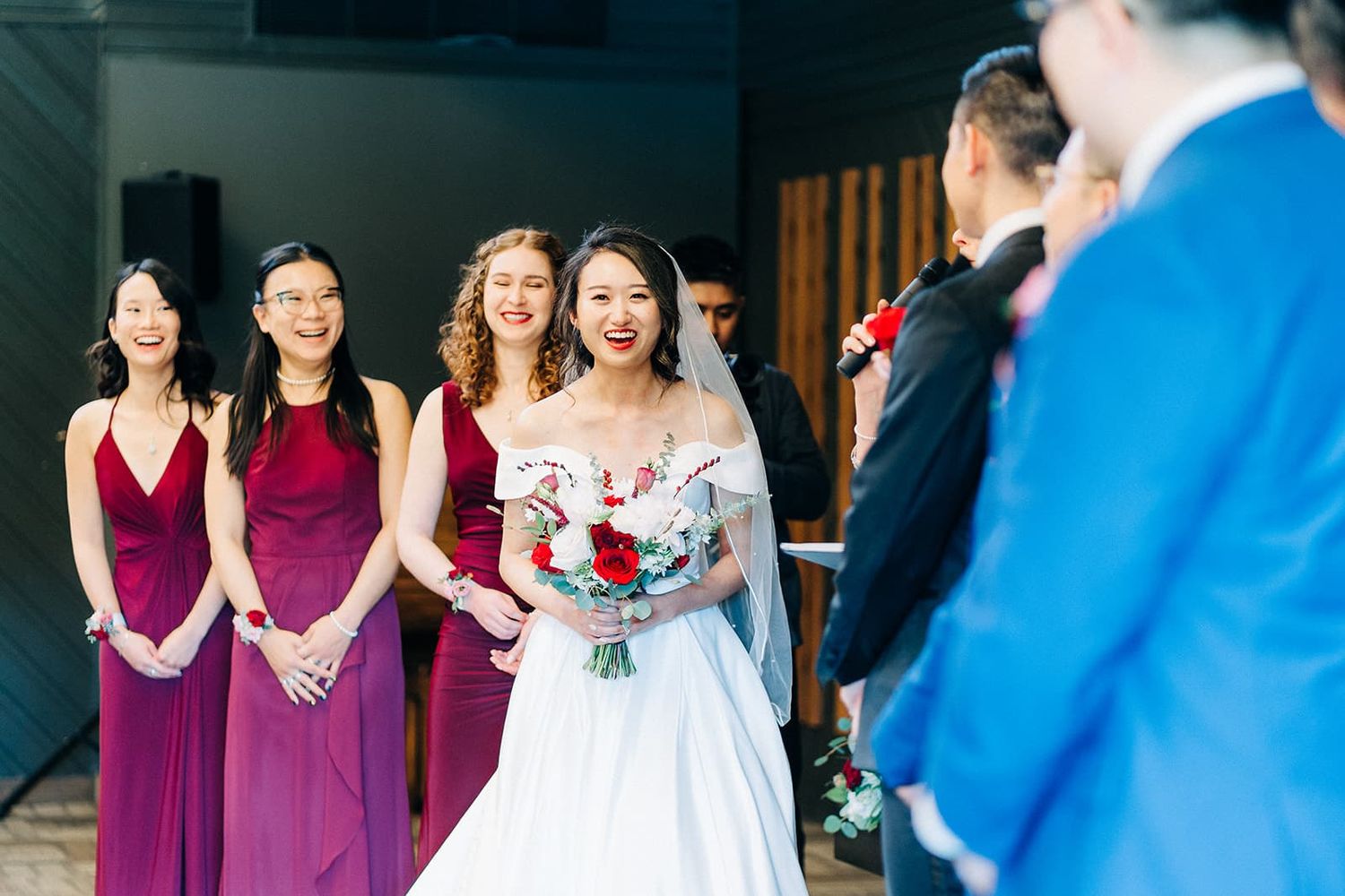 Bride in white gown holds red bouquet, smiling with bridesmaids in burgundy dresses at wedding ceremony.