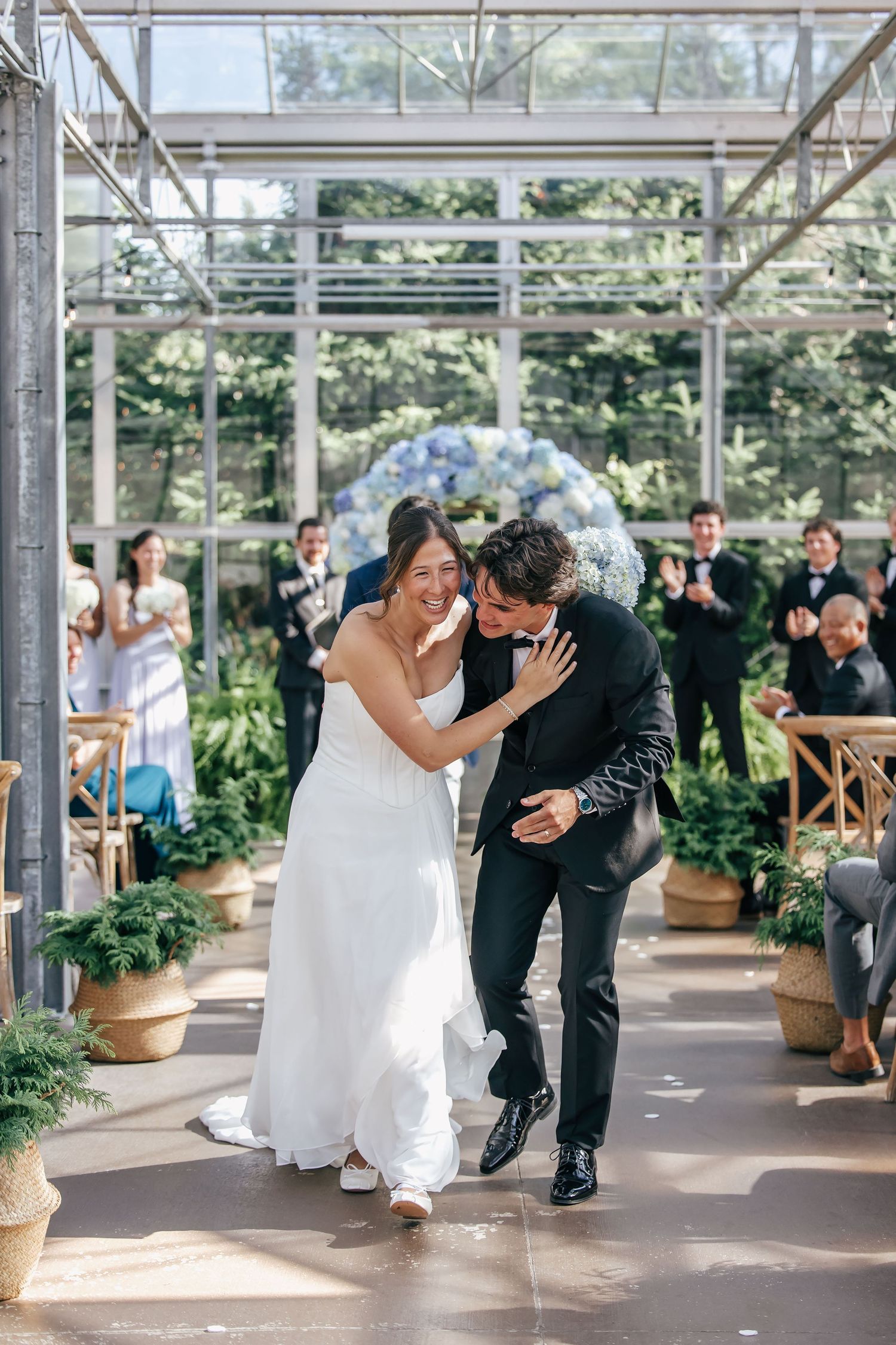 Joyful newlyweds walk down the aisle in a greenhouse wedding venue, surrounded by applauding guests and blue floral arch.