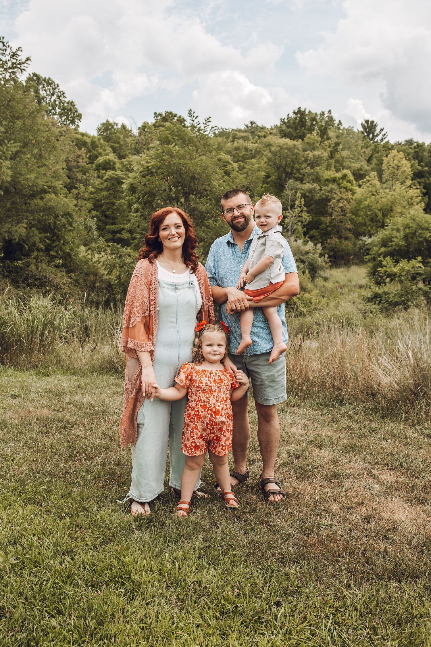 A family wearing coordinating orange and blue outfits poses together in a sunny meadow with green trees in background.