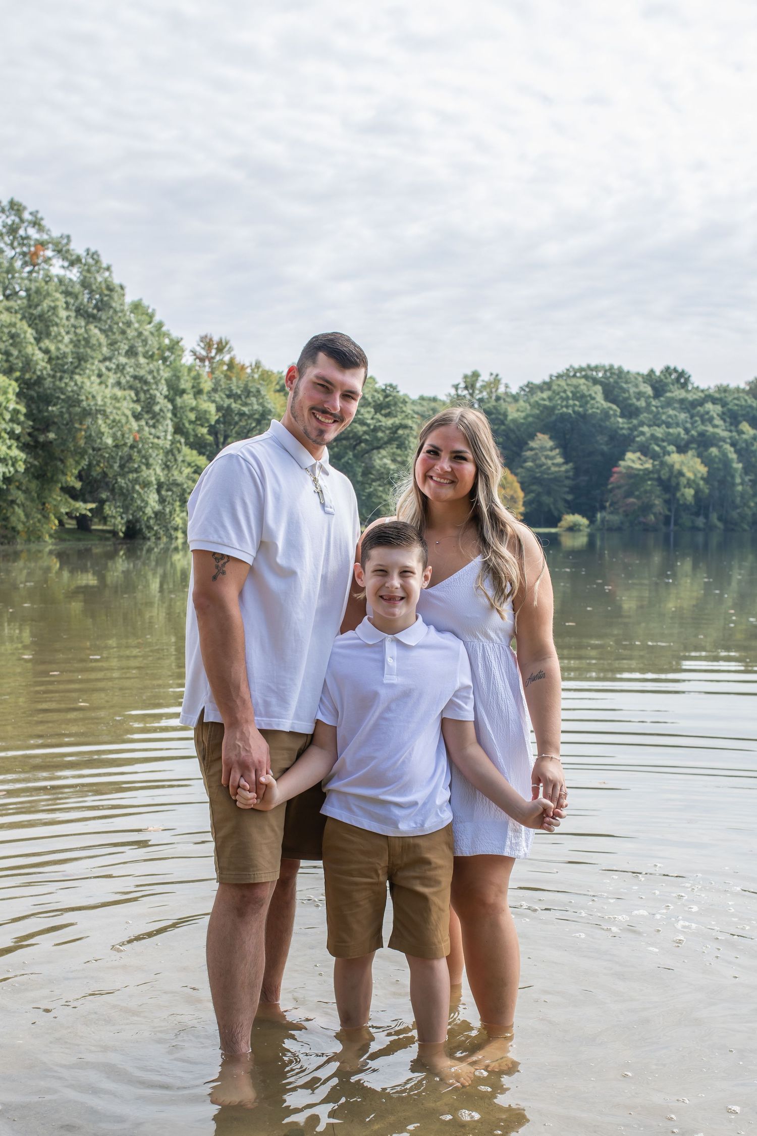 Family wearing white and khaki outfits stands together in shallow lake water on a cloudy summer day.