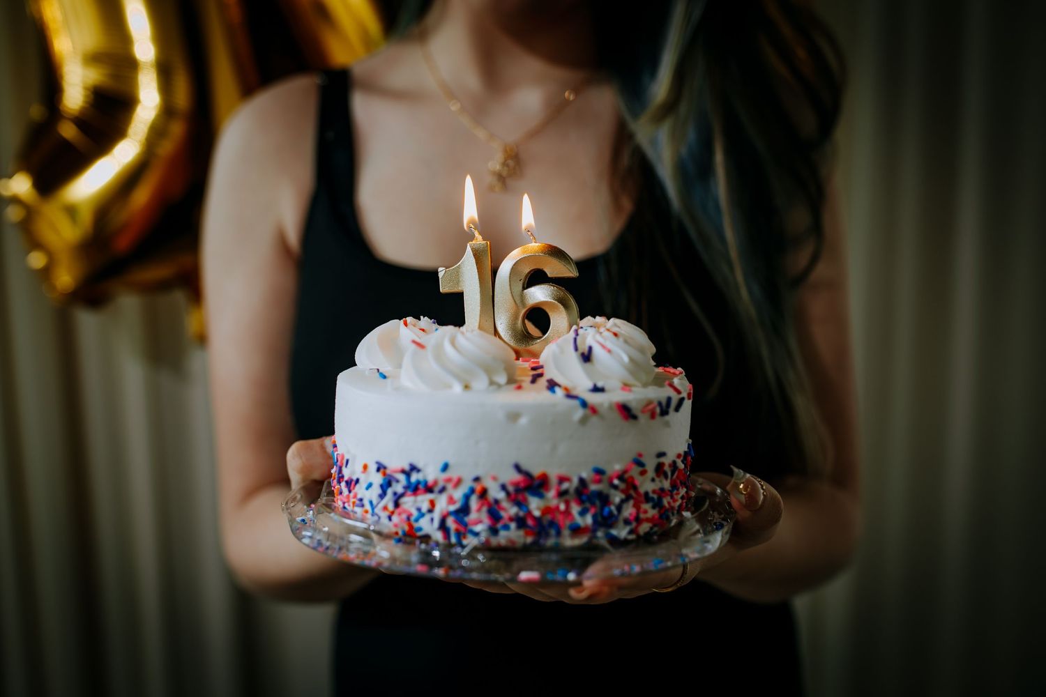 A young woman holds a white birthday cake with colorful sprinkles and lit number 16 candles at a sweet sixteen party.