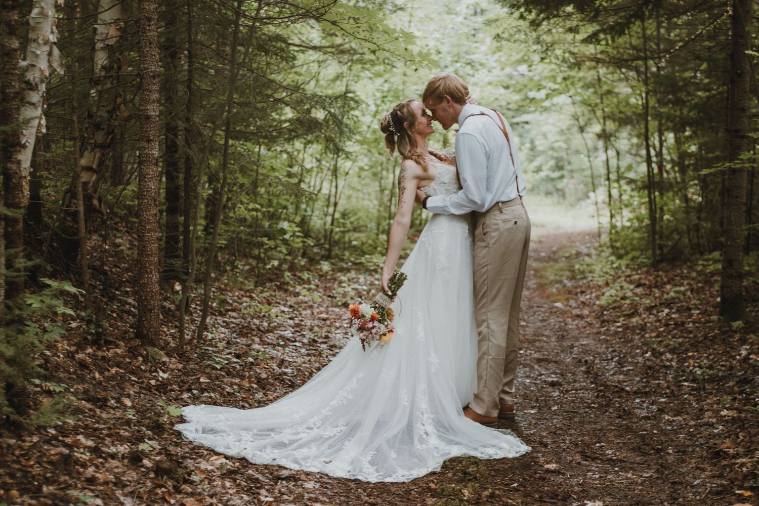 A couple shares a romantic moment on a wooded forest path, with the bride wearing a flowing white lace gown.