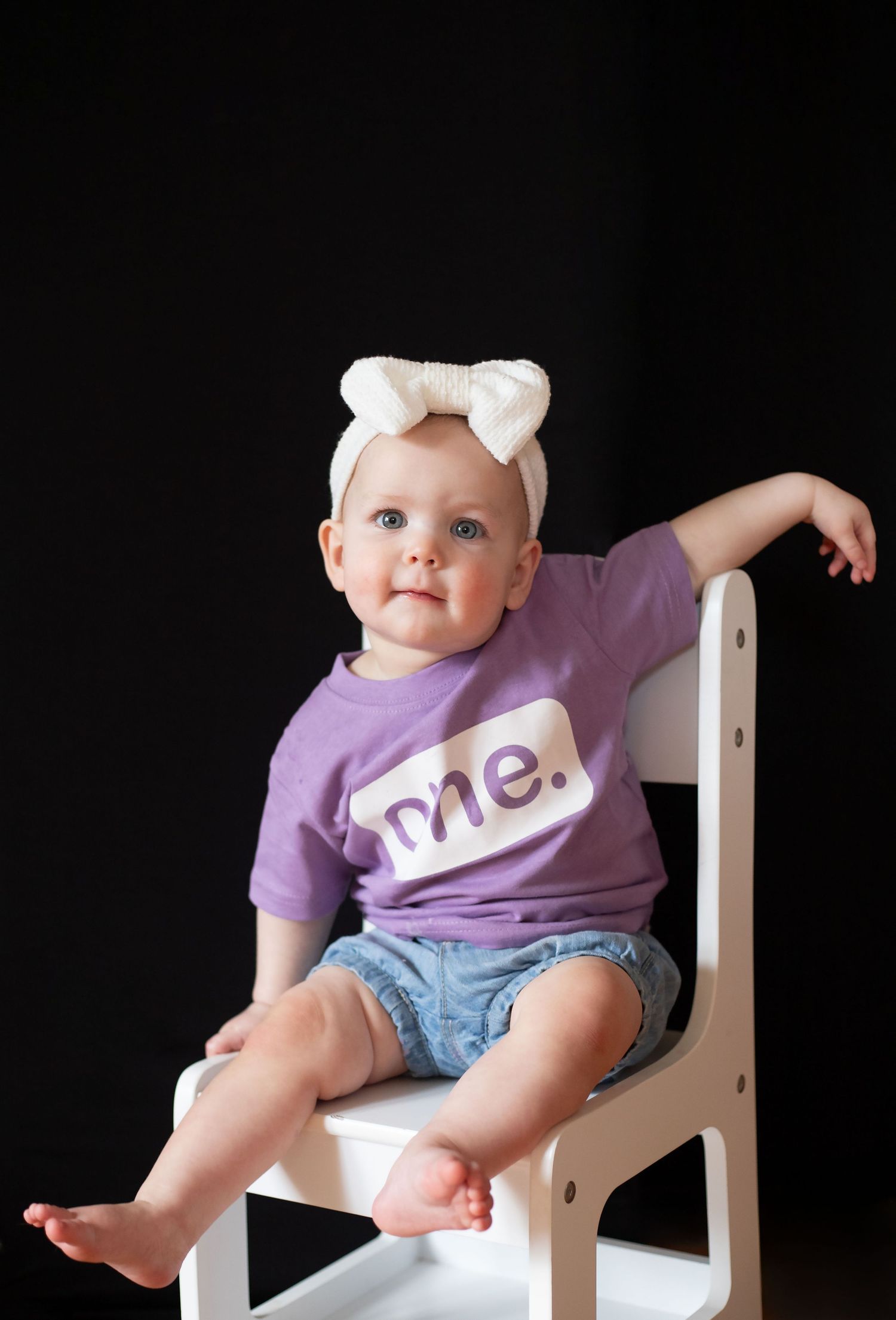 Baby girl in purple 'me.' shirt and white bow headband sitting on white chair against black background.