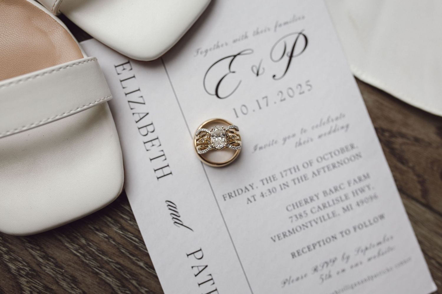 Wedding invitation for Elizabeth and Patrick on 10.17.2025 with gold diamond ring and white bridal heels on wooden surface.