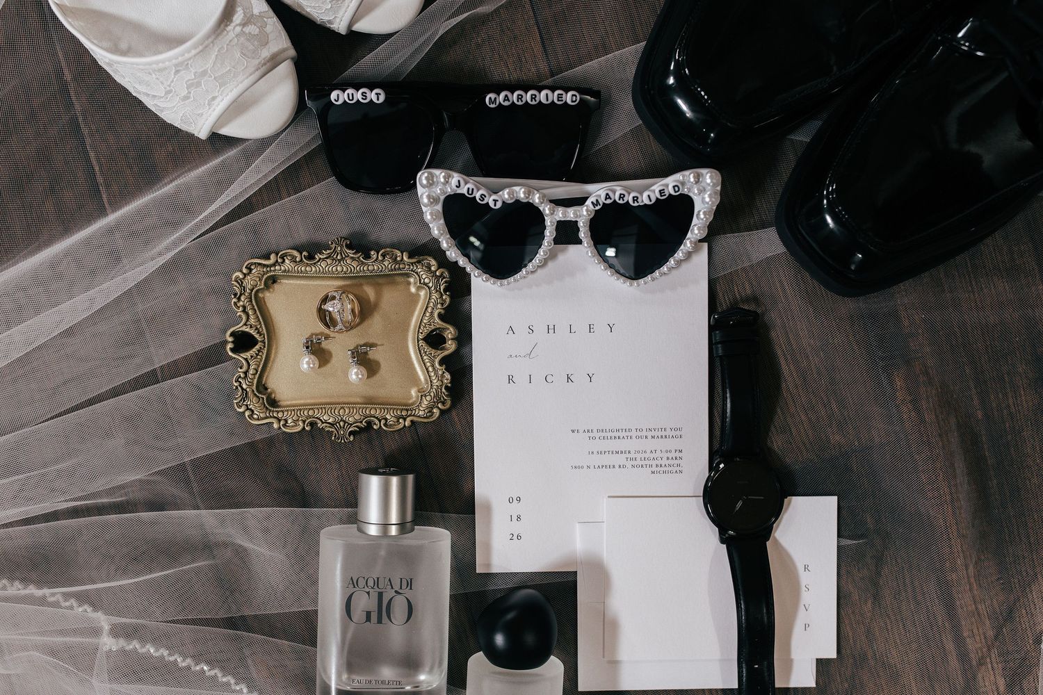 Elegant wedding flat lay featuring heart sunglasses, Armani Gio perfume, gold ring dish, black watch, and wedding invitation.