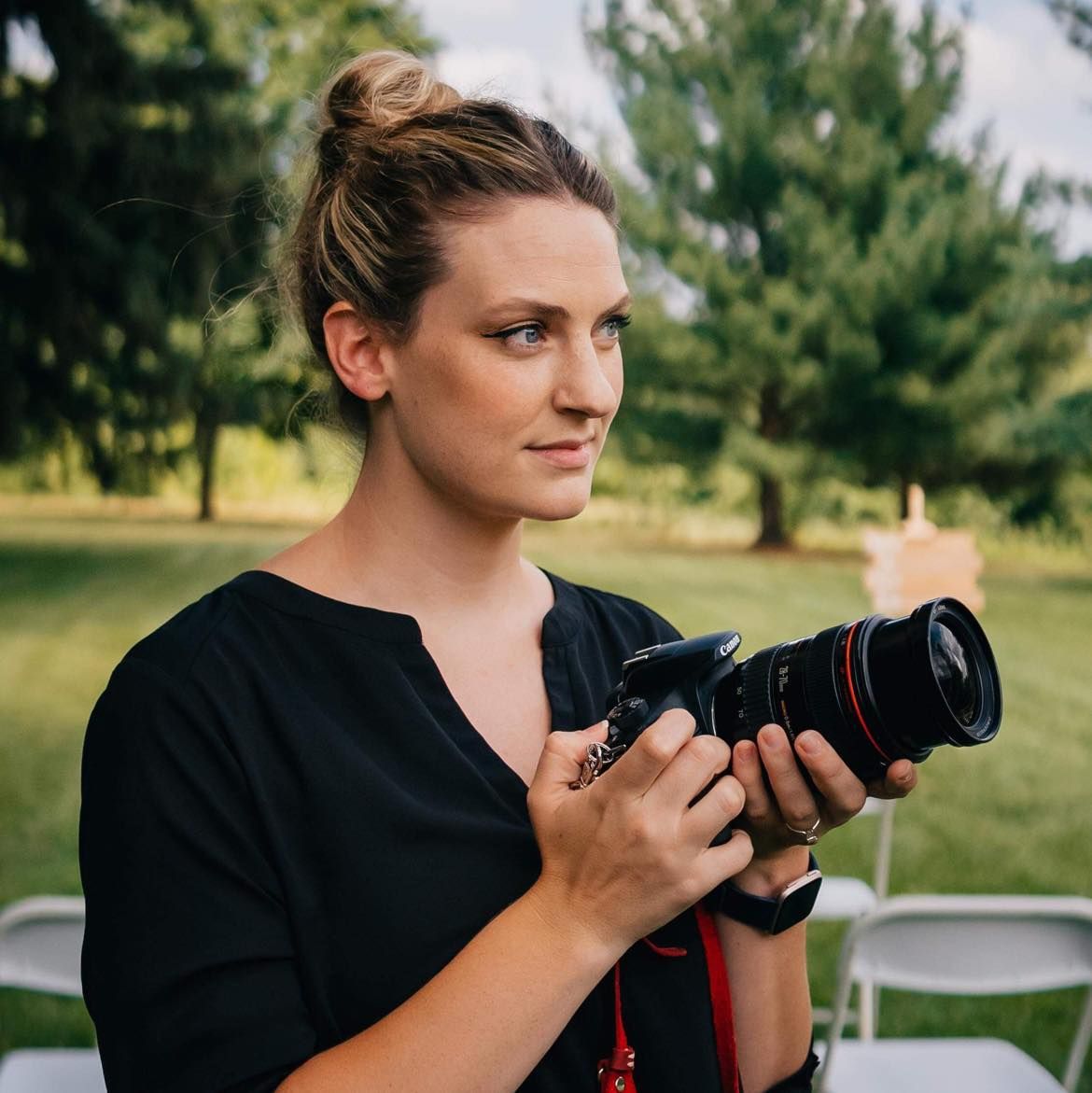 A female photographer in a black top holds a Canon DSLR camera outdoors at a green park setting.