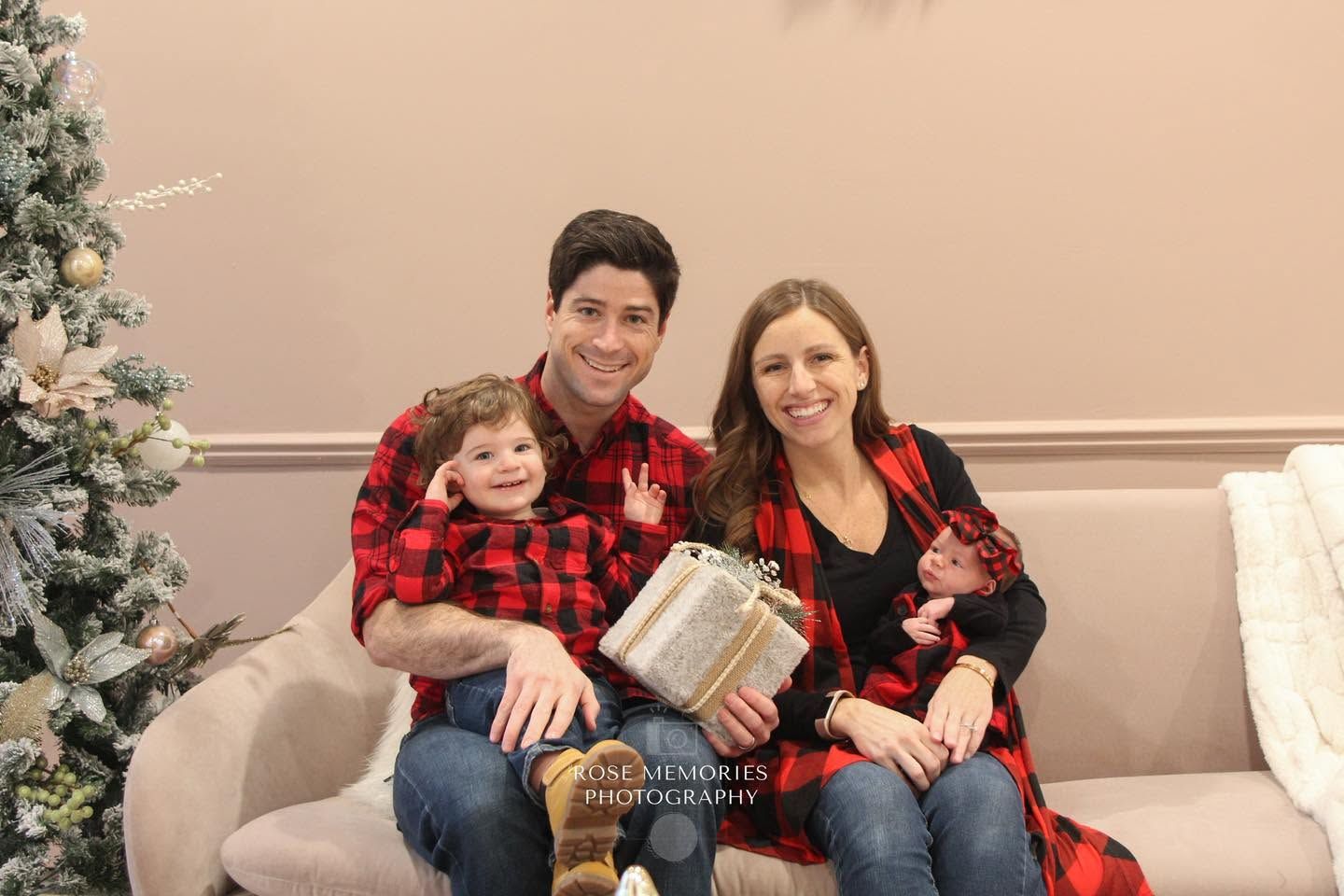 Family of three in matching red plaid outfits posing with a gift near a Christmas tree for a holiday portrait.