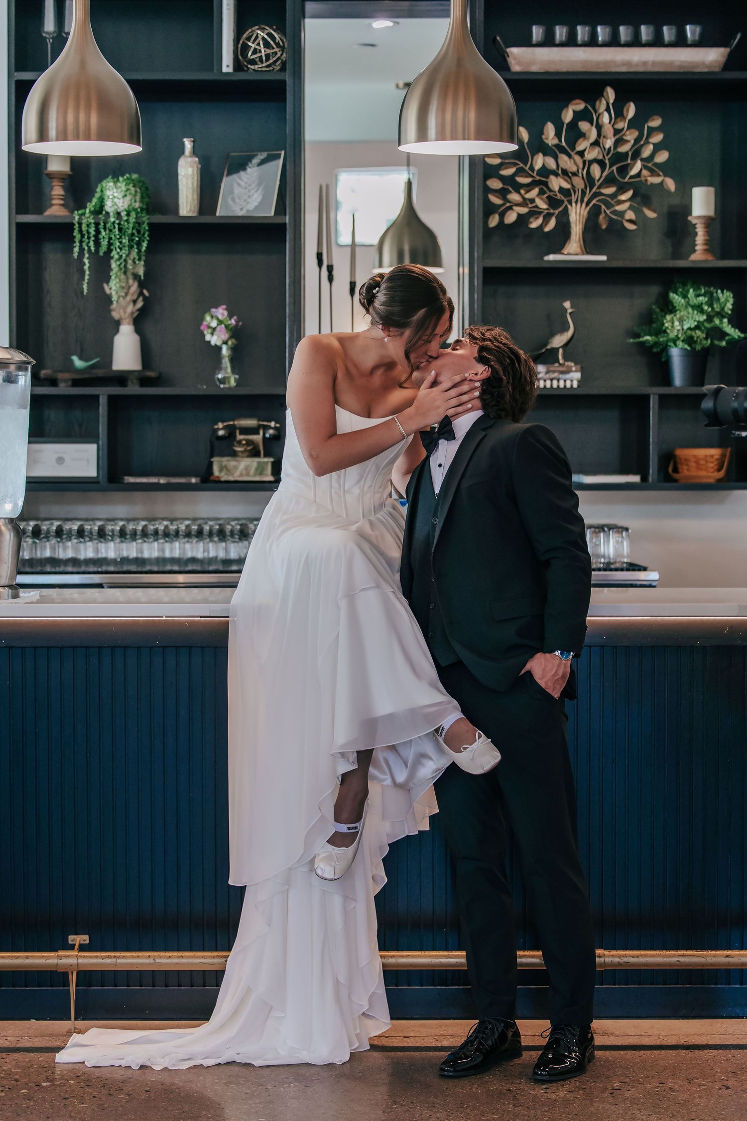 Bride and groom sharing a romantic kiss at a stylish bar, bride lifted in groom's arms, both in elegant wedding attire.