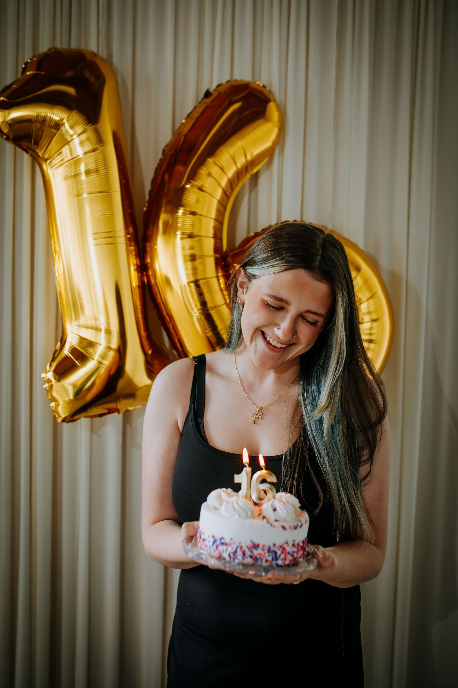 Young woman smiling while holding a 16th birthday cake with lit candles, gold number balloons in background.