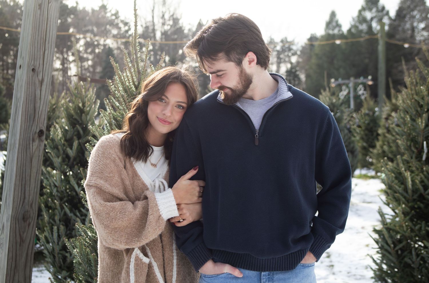 Couple embracing at a Christmas tree farm in winter, woman in tan wrap holding a mug, man in navy pullover.