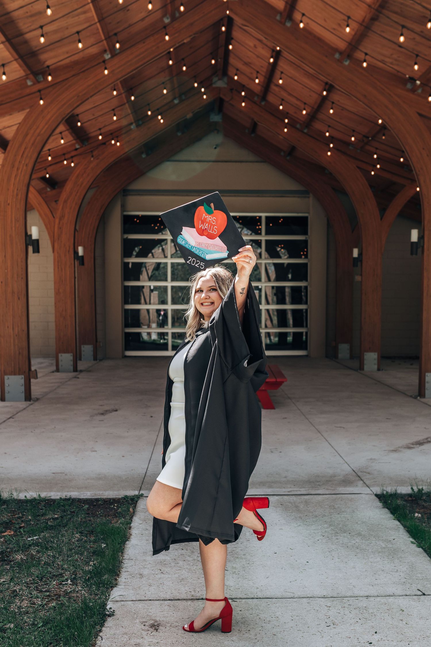 Smiling graduate in black gown holds decorated cap under wooden arch with string lights.