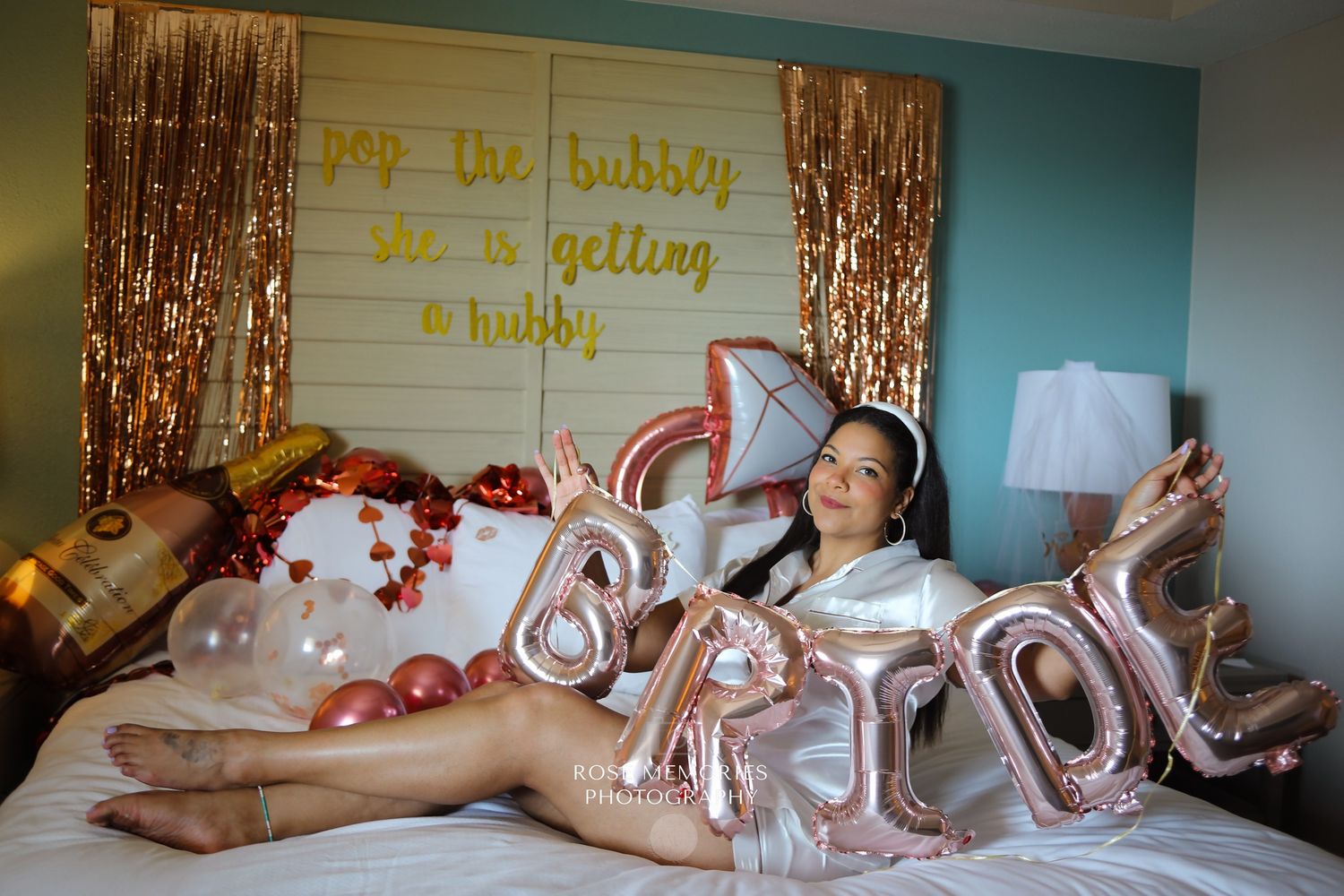 Woman posing on bed with rose gold BRIDE balloons, confetti, and pop the bubbly she is getting a hubby sign.