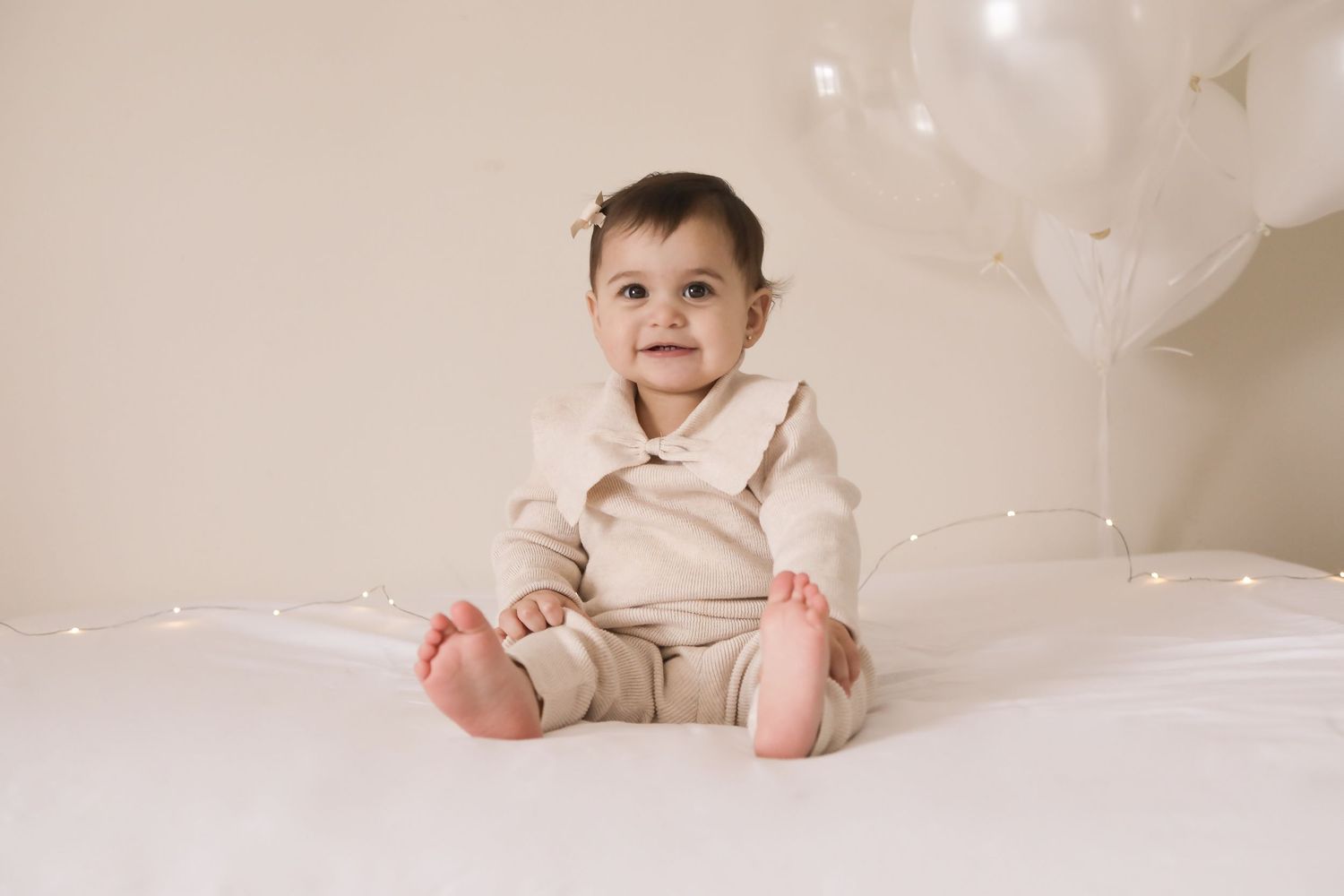 Smiling baby in beige outfit sits on white bed surrounded by white balloons and fairy lights.