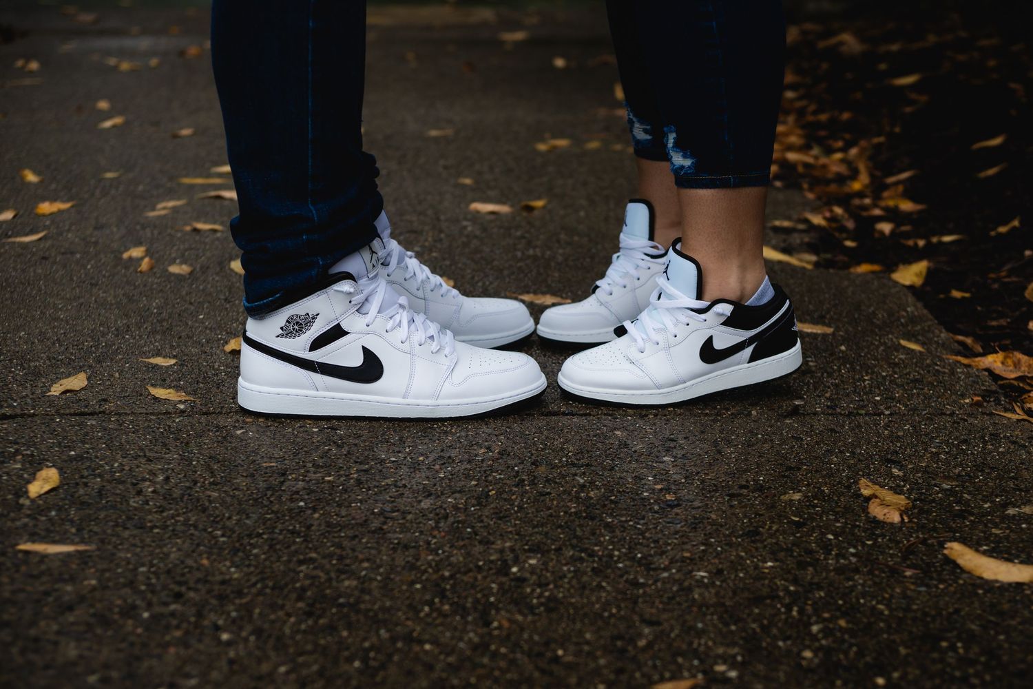 Two pairs of matching white Nike sneakers facing each other on fallen autumn leaves.