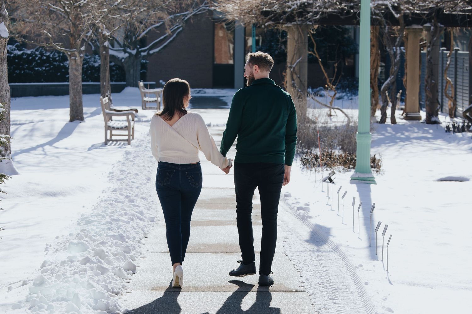 A couple walks hand in hand down a snowy path lined with bare winter trees.