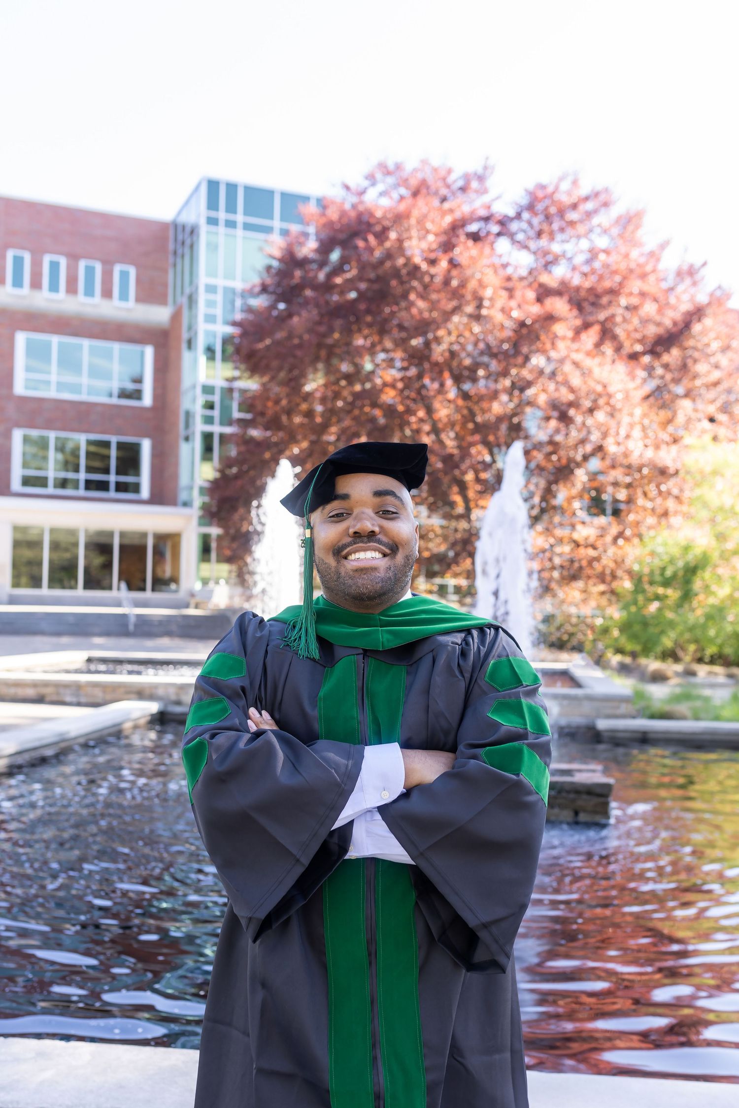 A graduate in green and gray academic regalia poses joyfully outdoors near a campus building.
