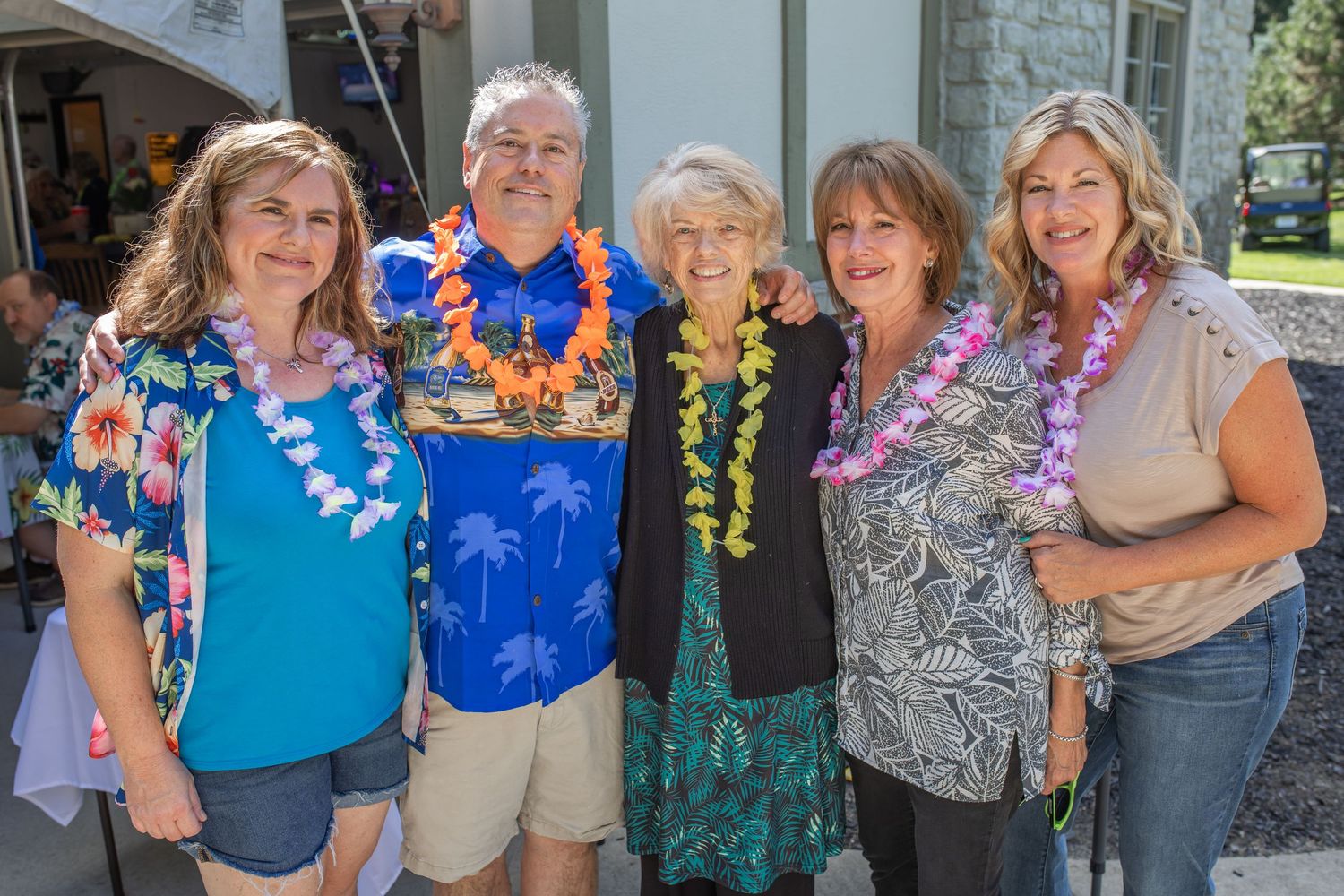 Five people wearing colorful Hawaiian leis smile together at an outdoor luau party on a sunny day.