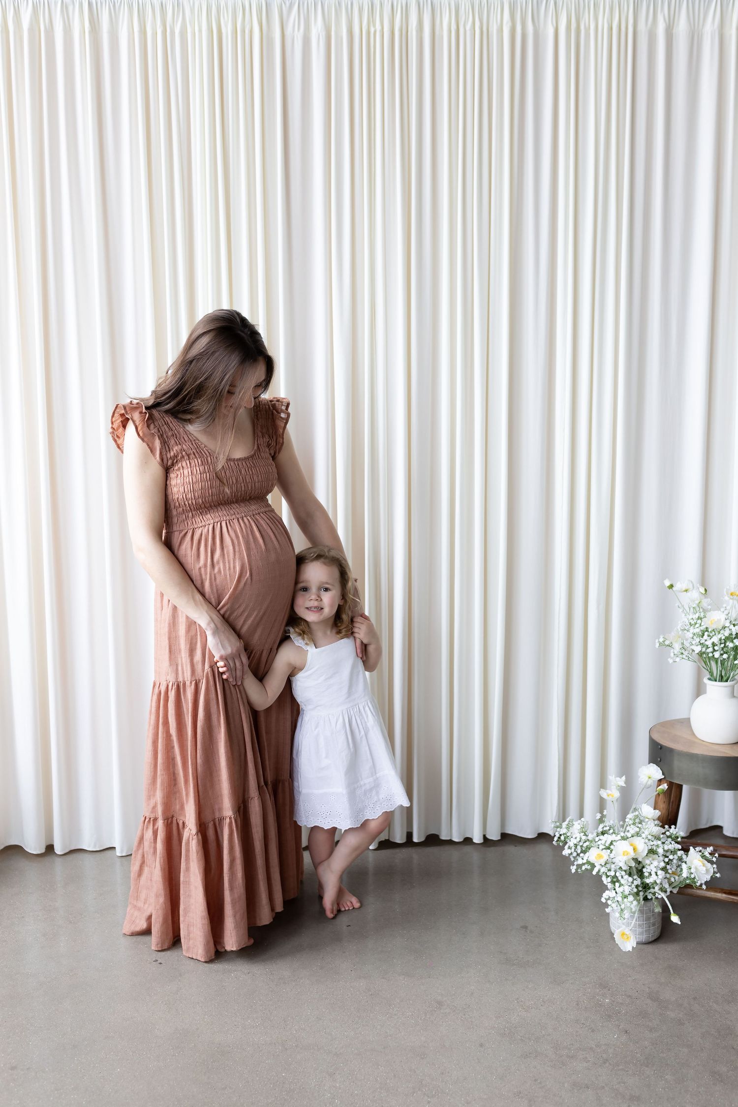 Pregnant woman in pink dress holds hands with toddler girl in white dress, standing before cream curtains.