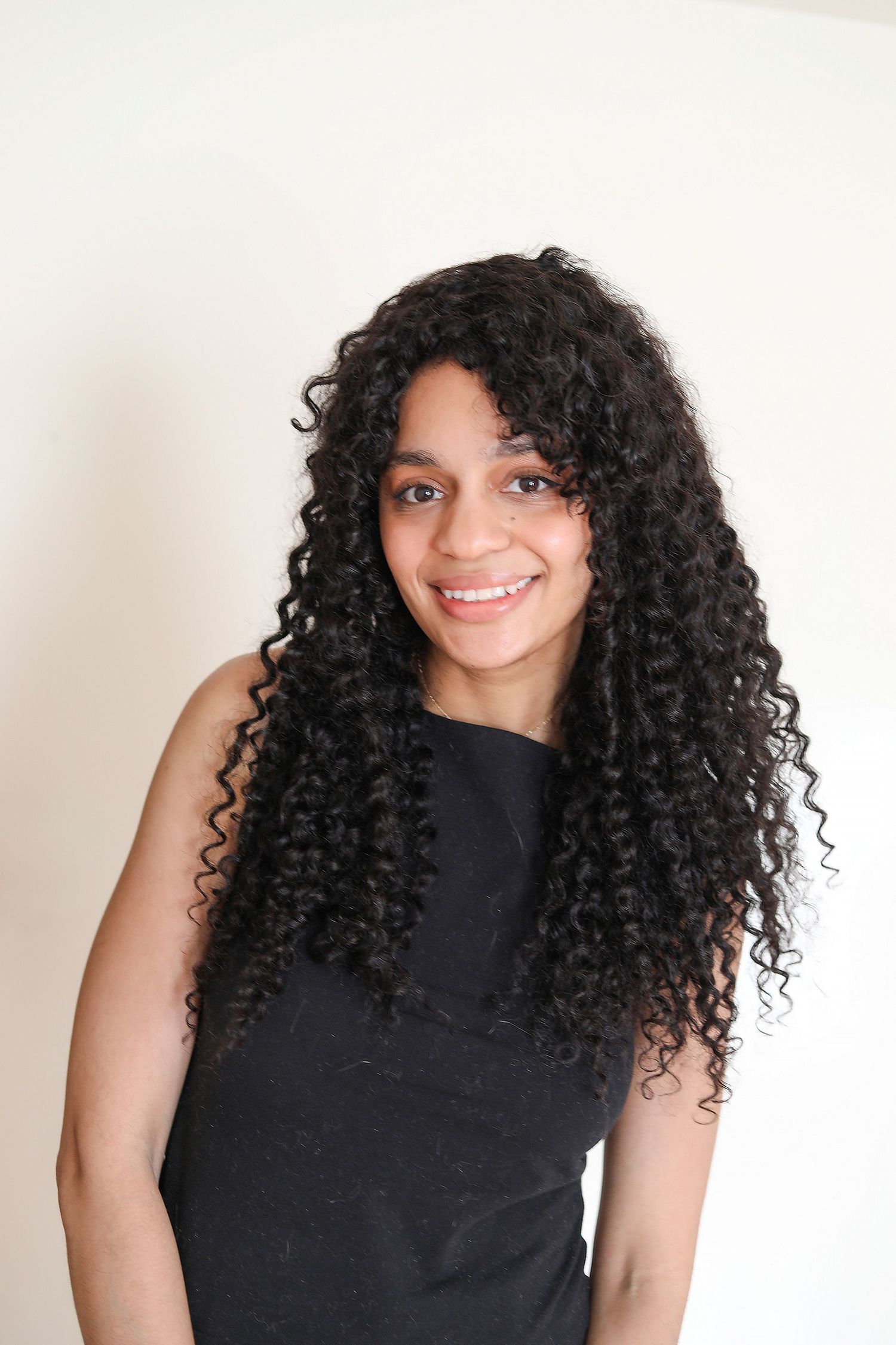 Young woman with long curly black hair smiling, wearing a black sleeveless top against a white background.