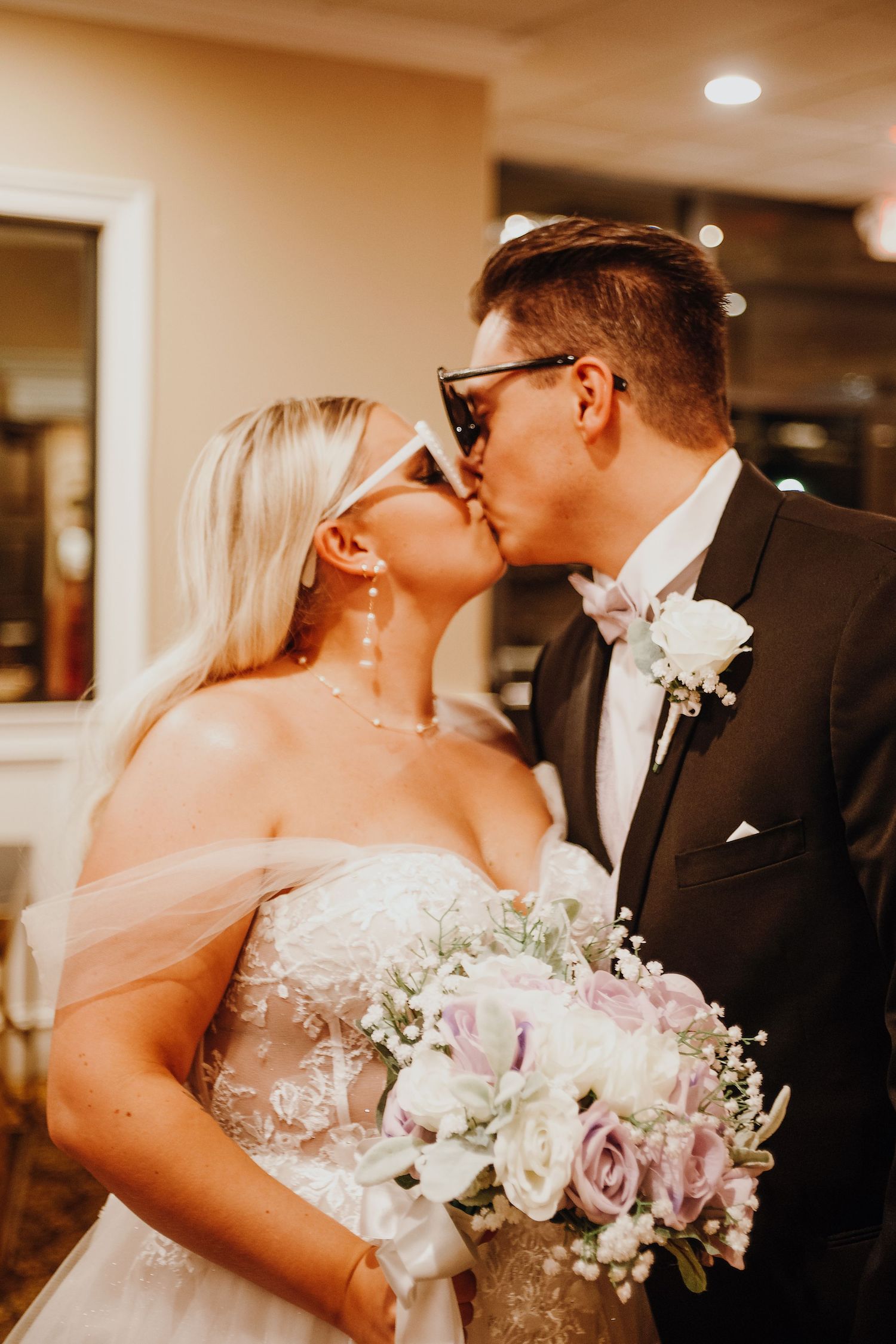 Bride and groom sharing a kiss at their wedding, bride holding a bouquet of pink and white roses, both wearing sunglasses.