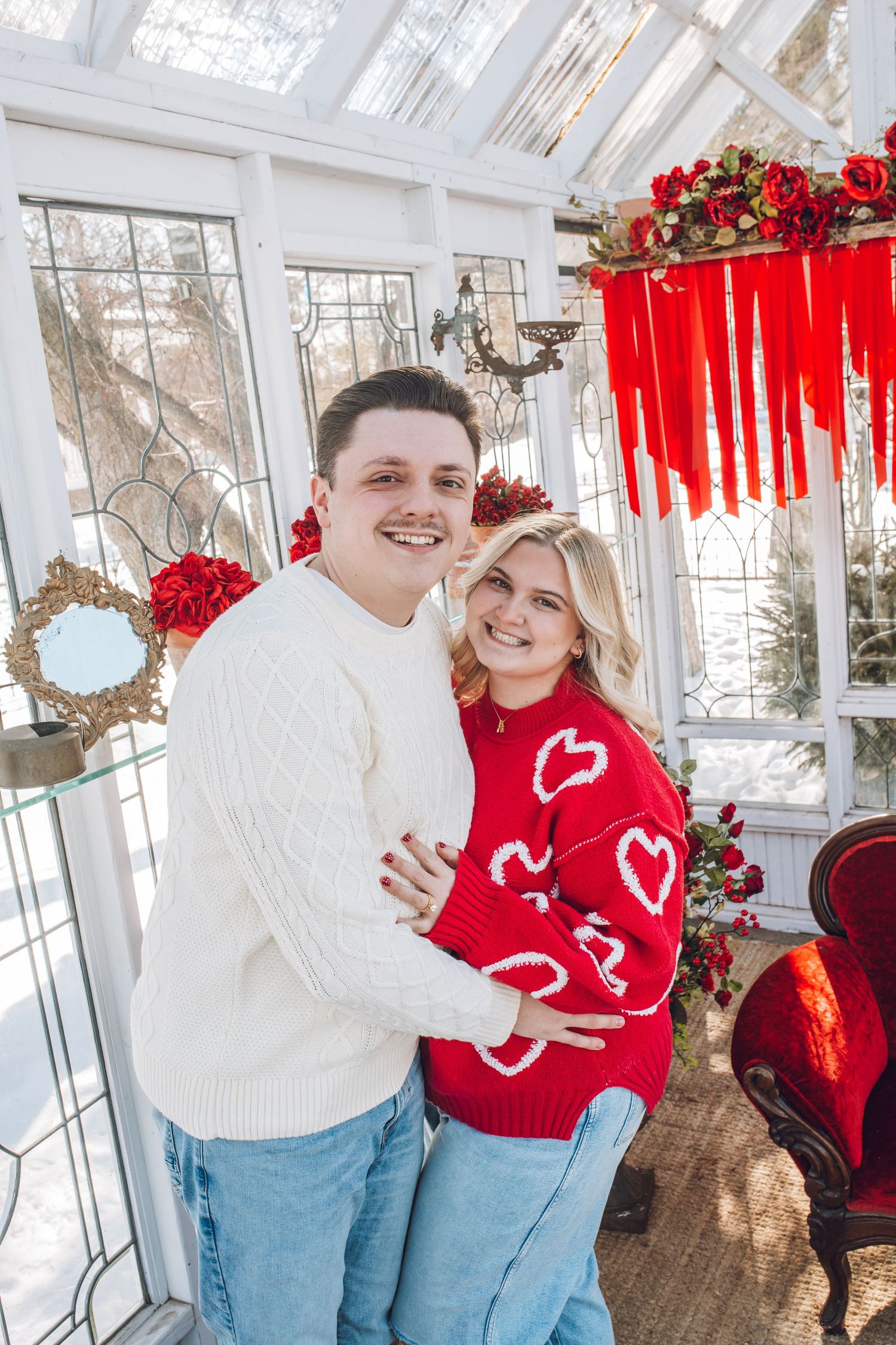 A couple in cozy knit sweaters embraces in a Valentine's Day decorated greenhouse with red roses and heart accents.