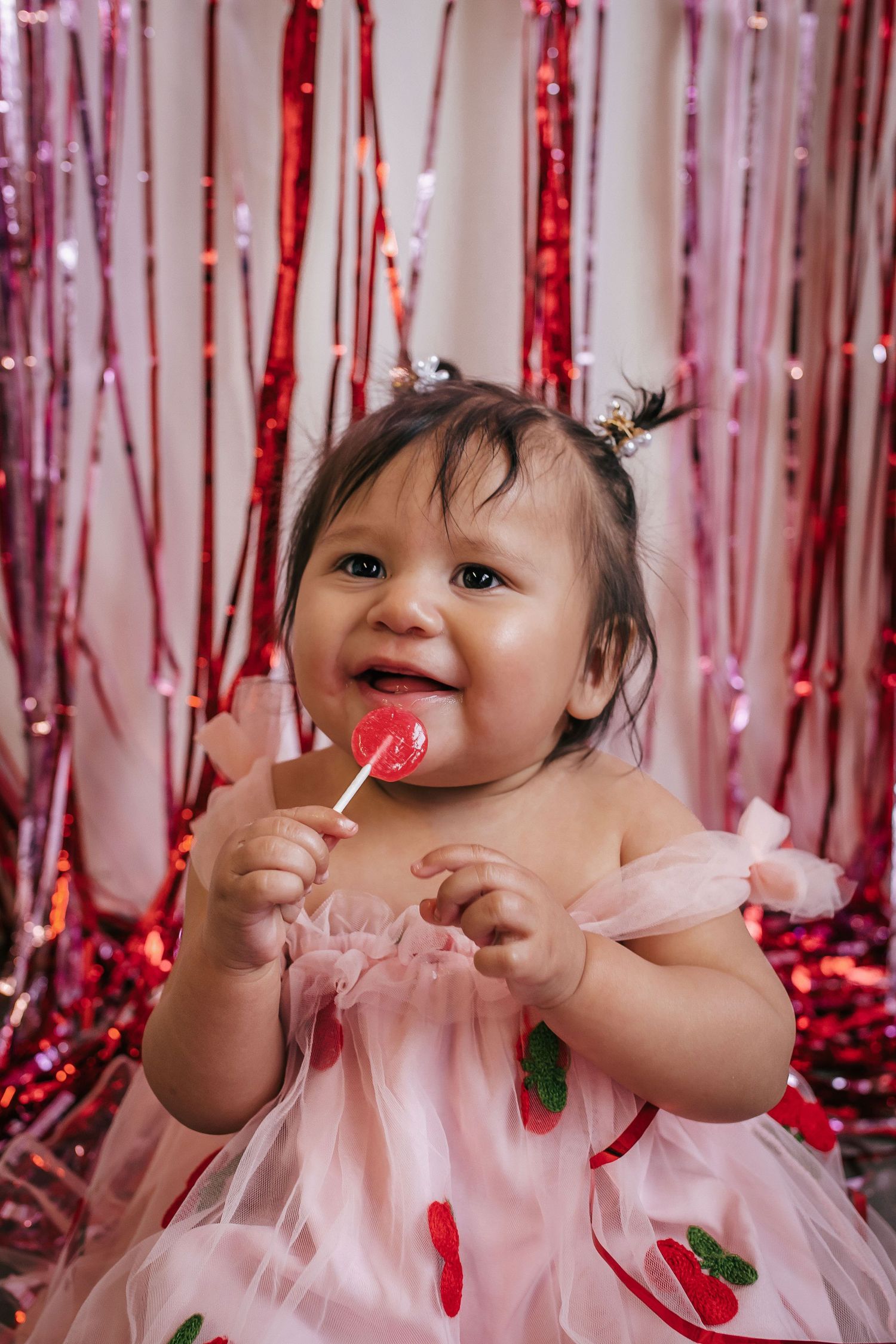 A happy baby girl in a pink tulle dress licks a heart-shaped lollipop against a shiny red tinsel backdrop.