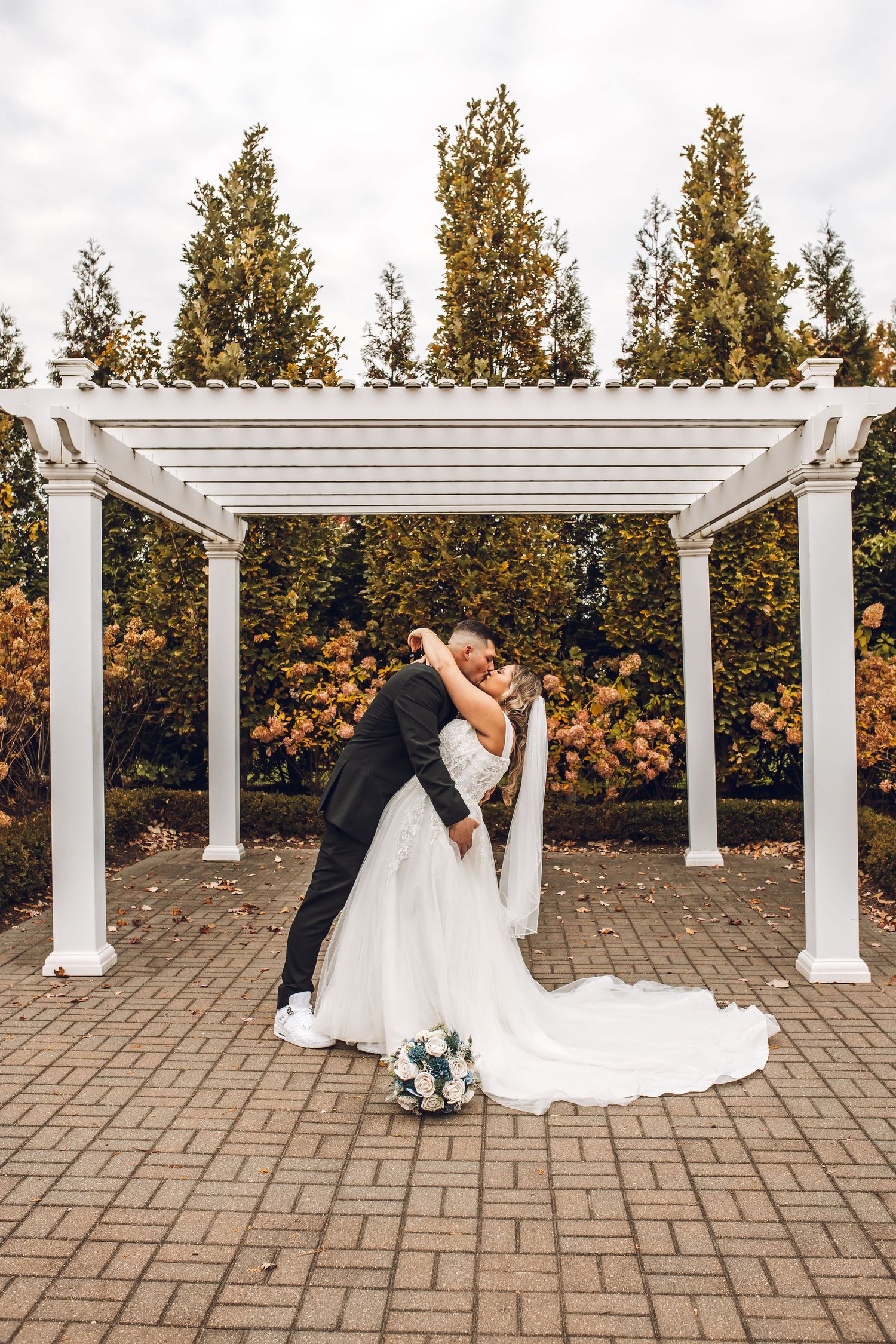 A romantic wedding couple shares an intimate moment under a white pergola on a brick patio during autumn.