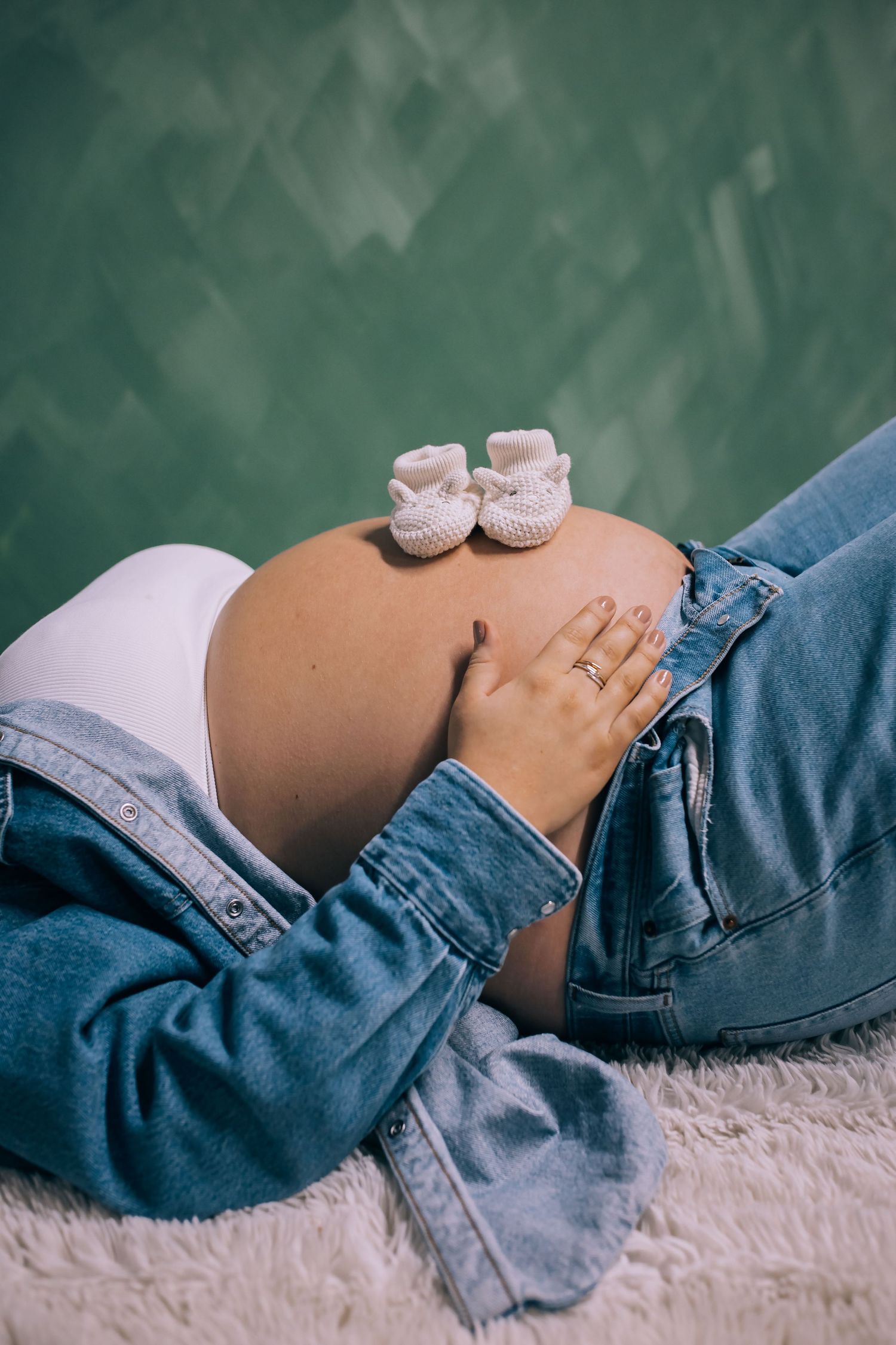 Pregnant woman lying down in denim jacket, cradling bare belly with tiny white knitted baby booties resting on top.