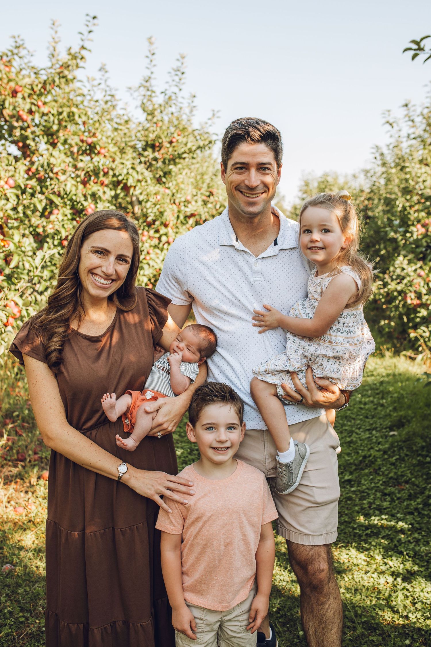 A family poses together outdoors in an orchard during golden hour wearing brown and white casual clothing.