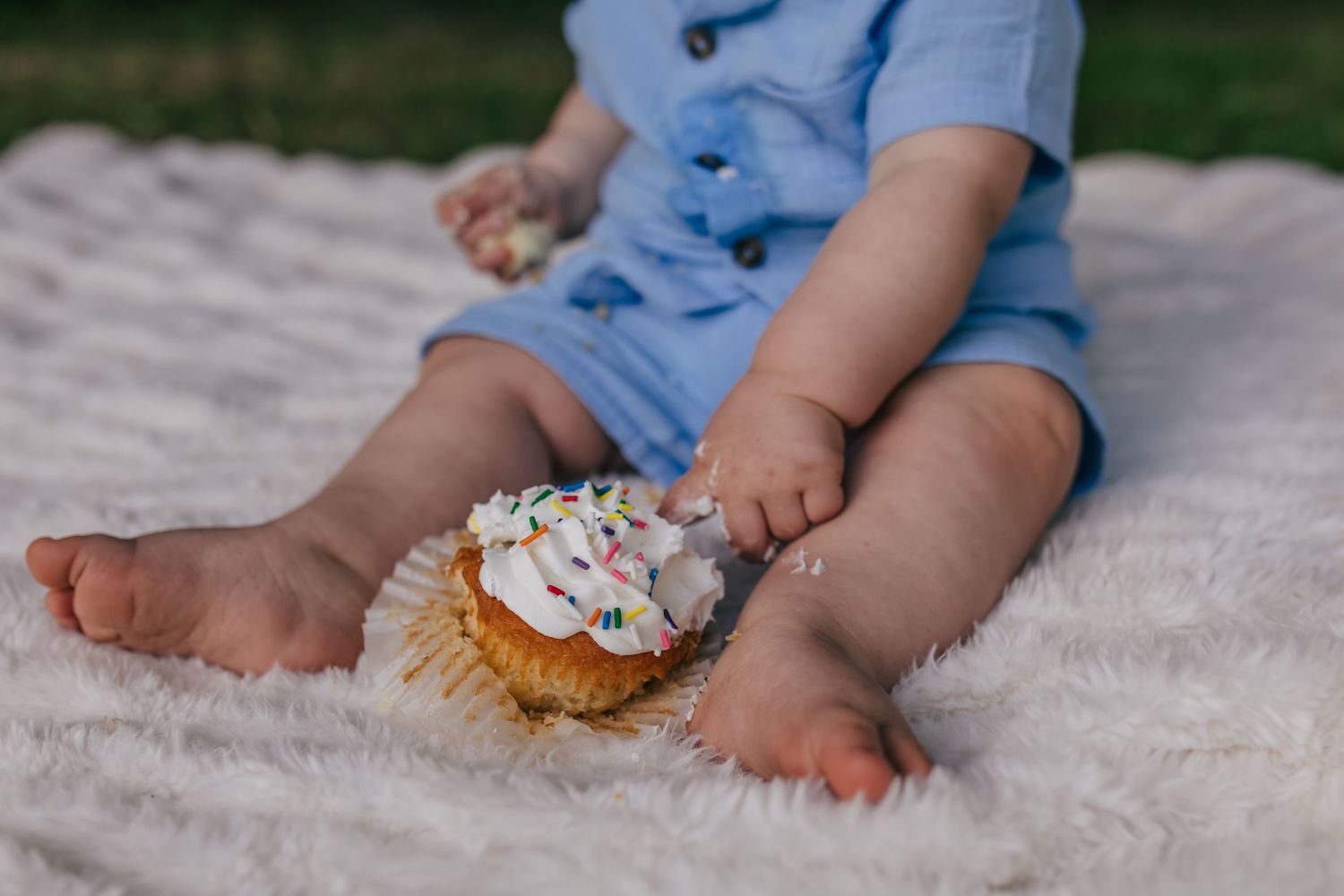 Baby in blue romper sitting on white blanket outdoors, touching a cupcake with white frosting and sprinkles.