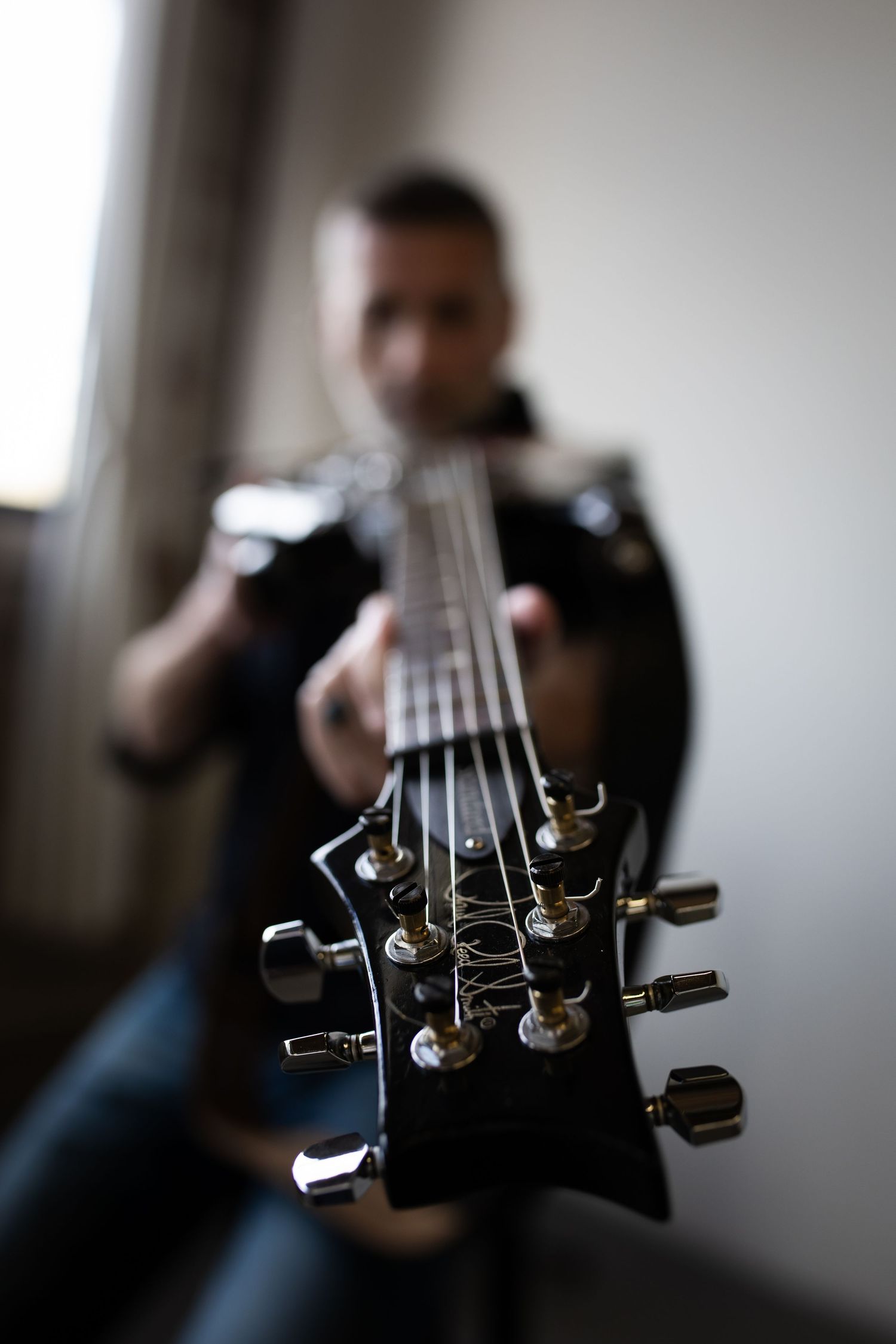 A musician holds a black electric guitar toward the camera, with the headstock and tuning pegs in sharp focus.