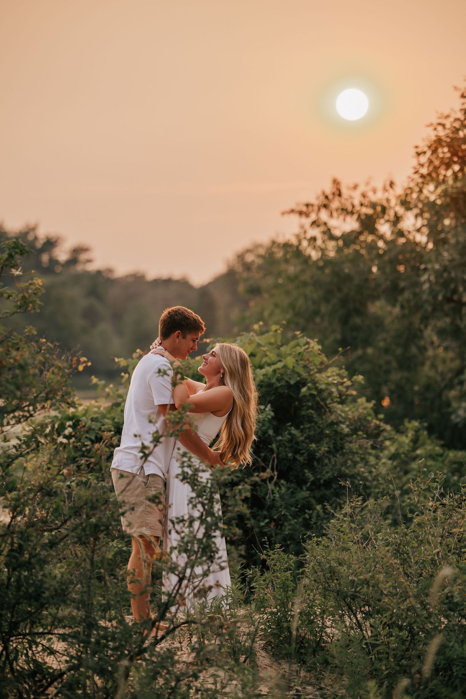 Couple embracing romantically in lush greenery during golden sunset, woman in white dress, warm glowing sky backdrop.