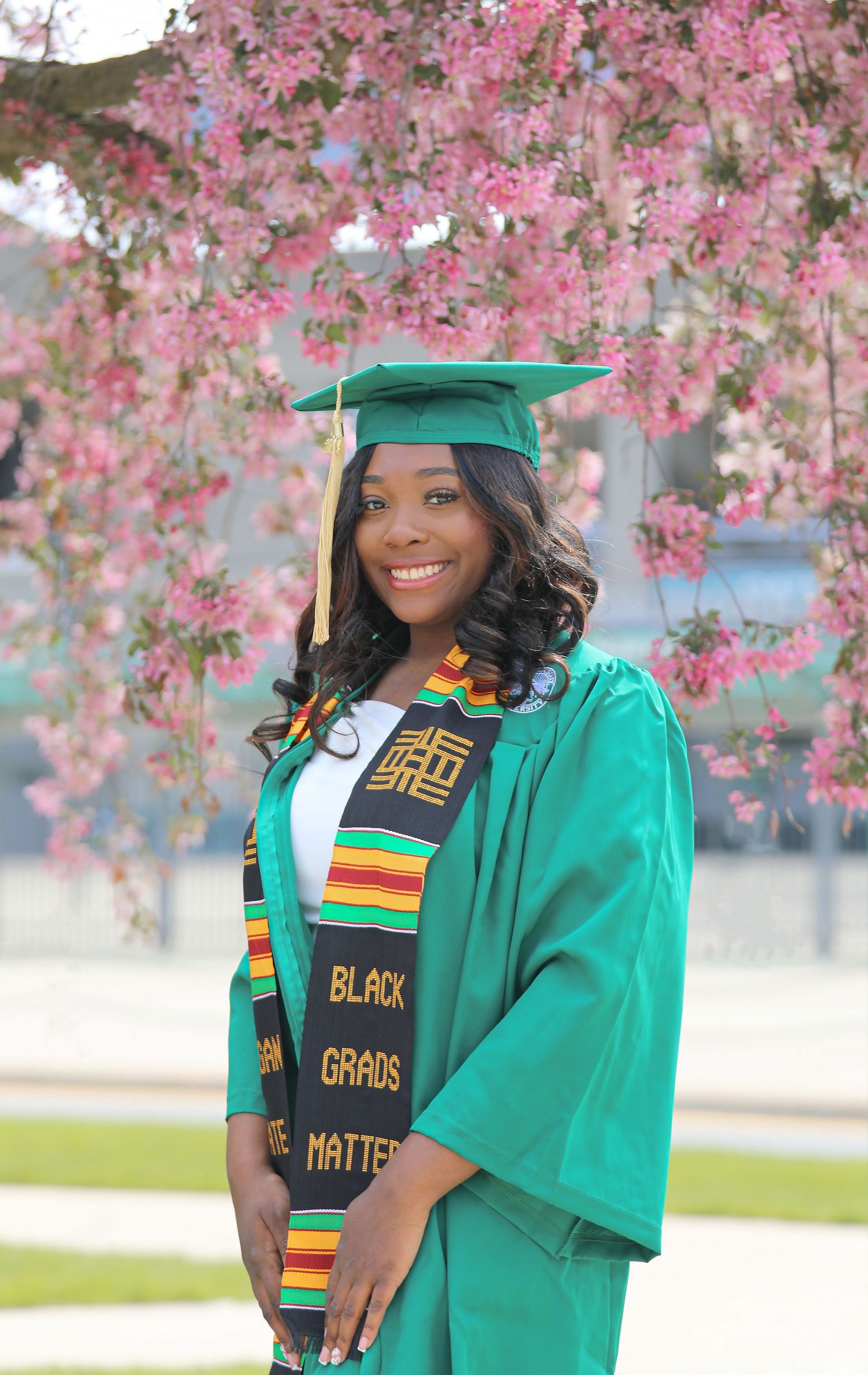 Young woman in green graduation cap and gown wearing a Black Grads Matter kente stole, posing under pink blossoms.
