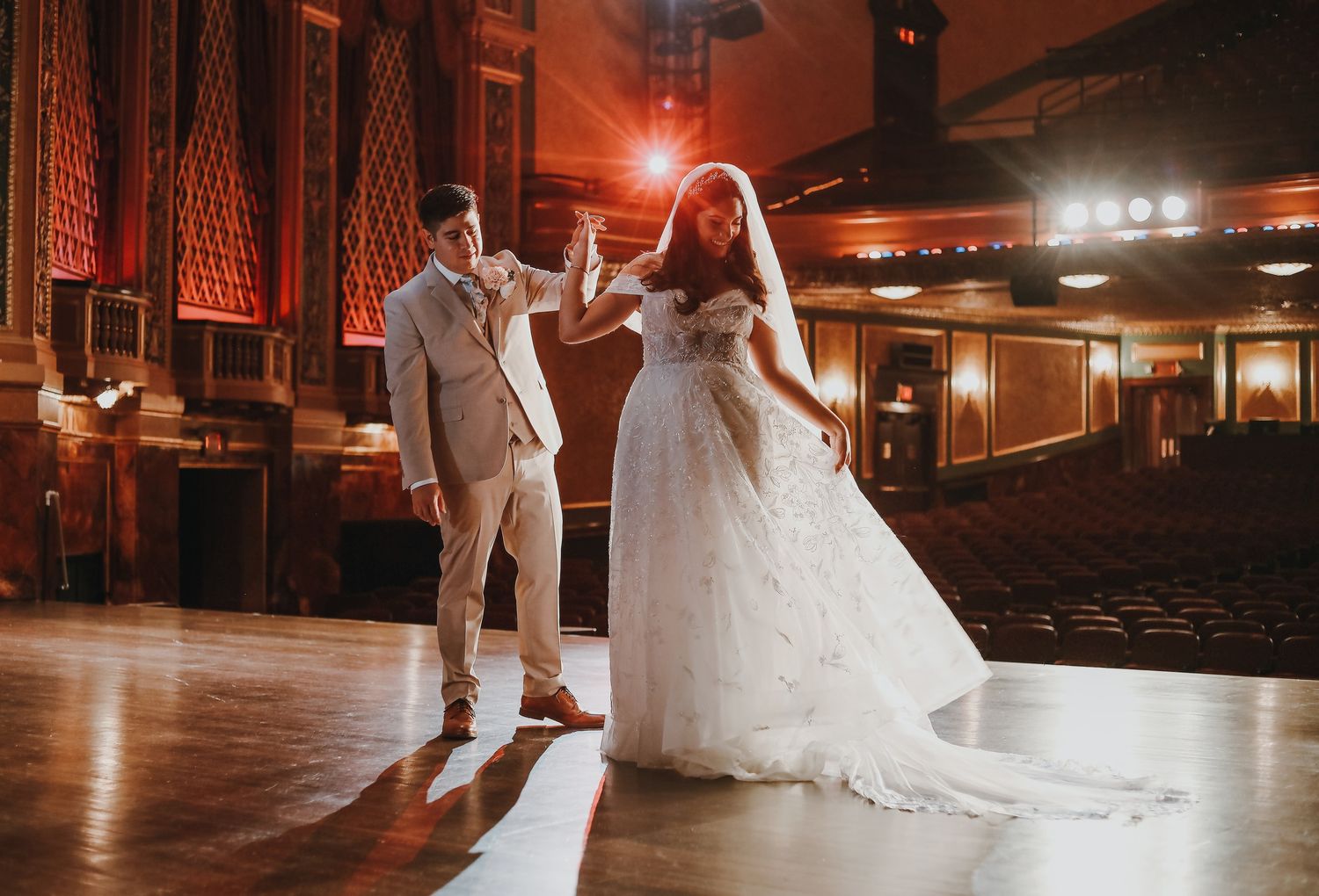 Bride in white gown and groom in tan suit share first dance in elegant theater venue with warm lighting.