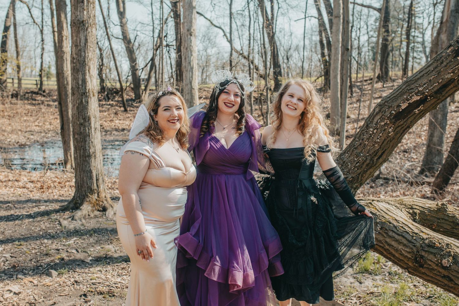 Three women in formal gowns pose outdoors in a wooded setting, wearing white, purple, and black dresses.