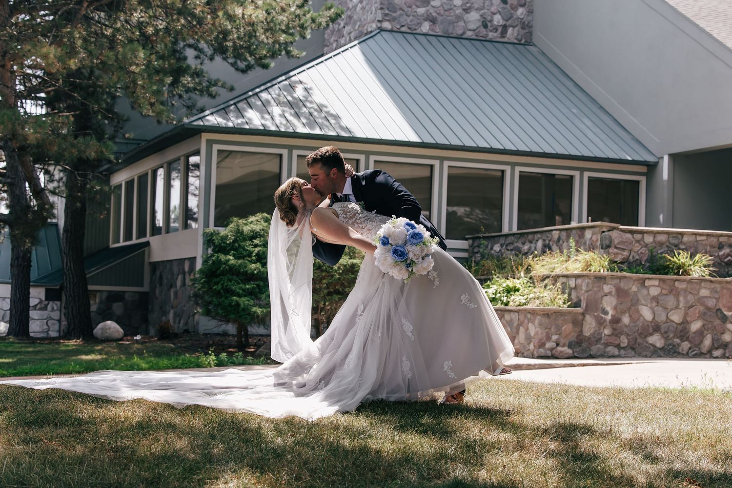 Bride and groom sharing a romantic dip kiss outdoors in front of a stone building on their wedding day.