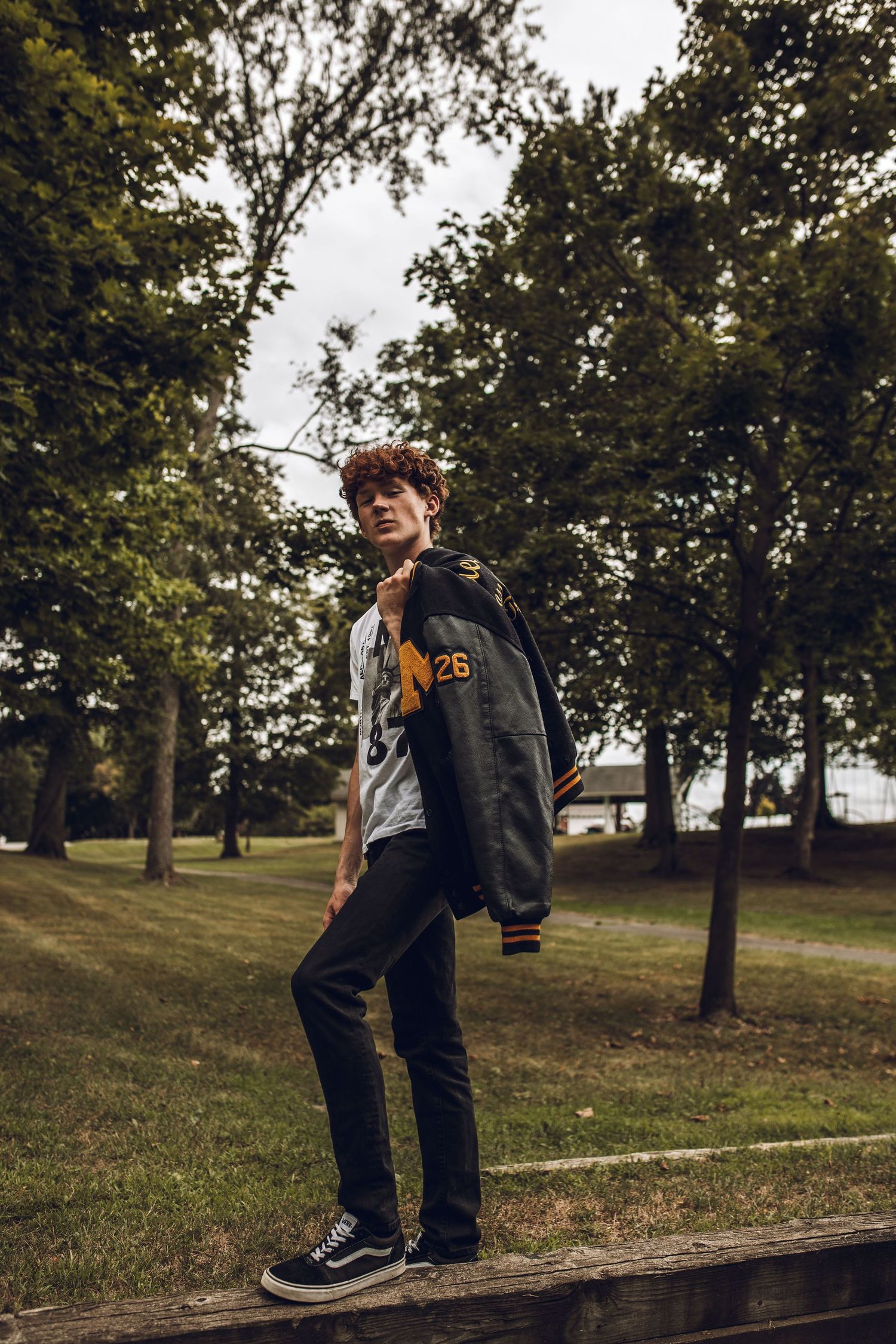 Young man in varsity jacket and Vans sneakers posing in a lush green park on a wooden beam.