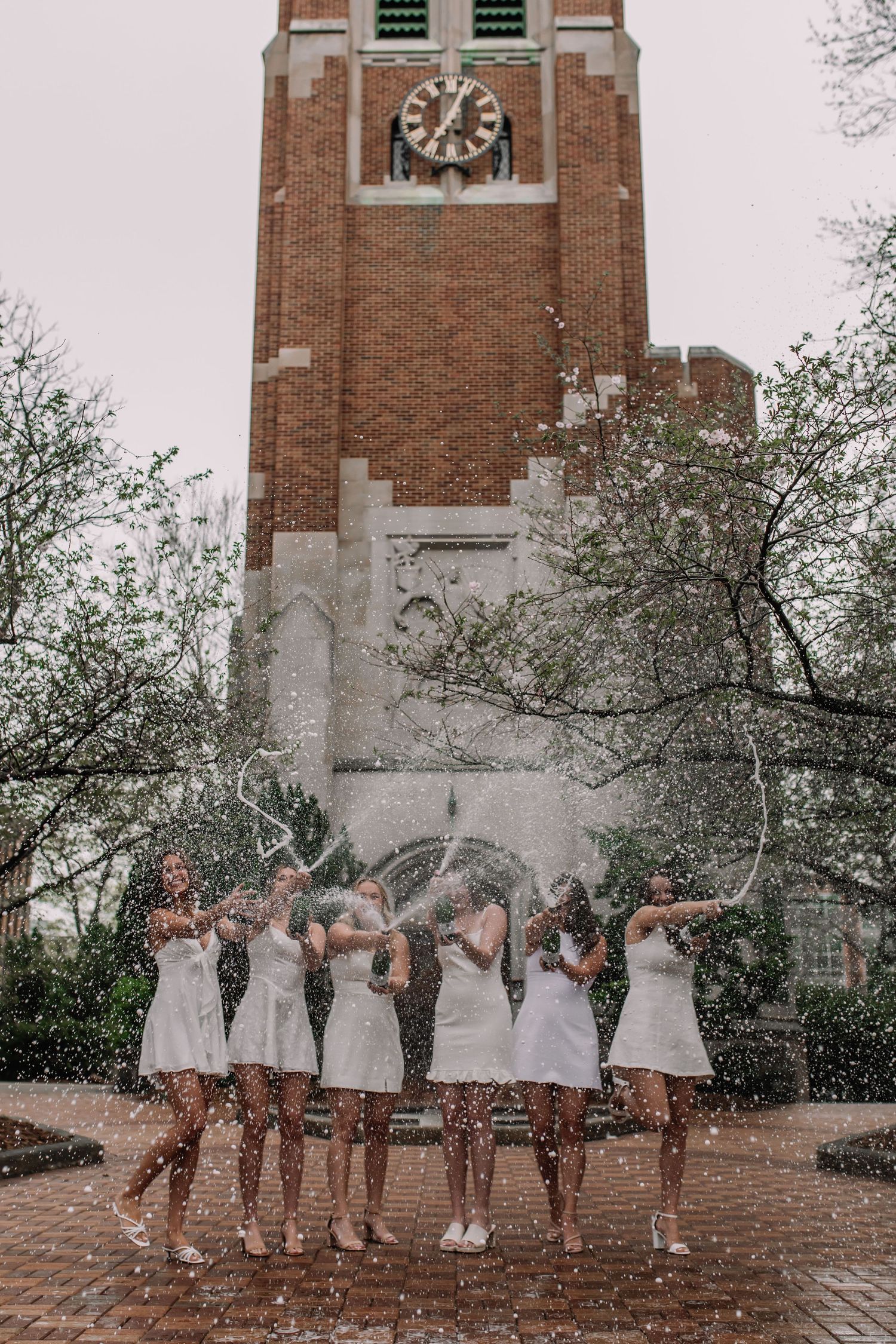 Six women in white dresses celebrate with champagne spray in front of a brick clock tower on a college campus.