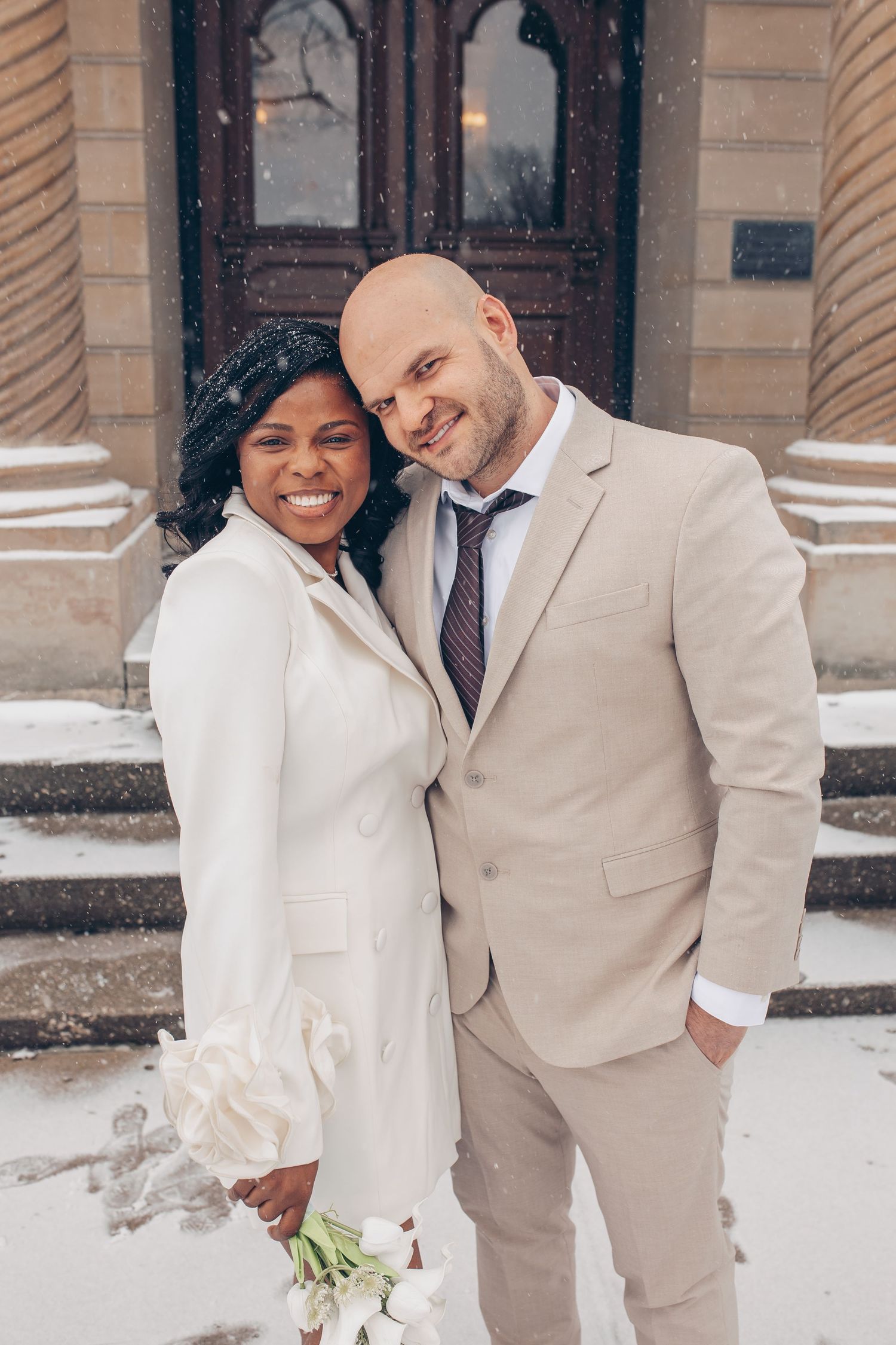 Interracial couple in wedding attire smiling together outside a snowy building, bride holding white tulips.
