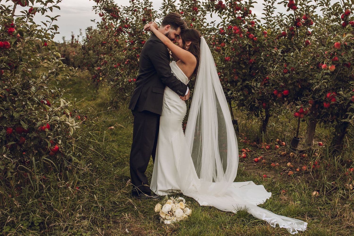 Bride and groom share romantic kiss in apple orchard, bride wearing flowing white gown and long veil.
