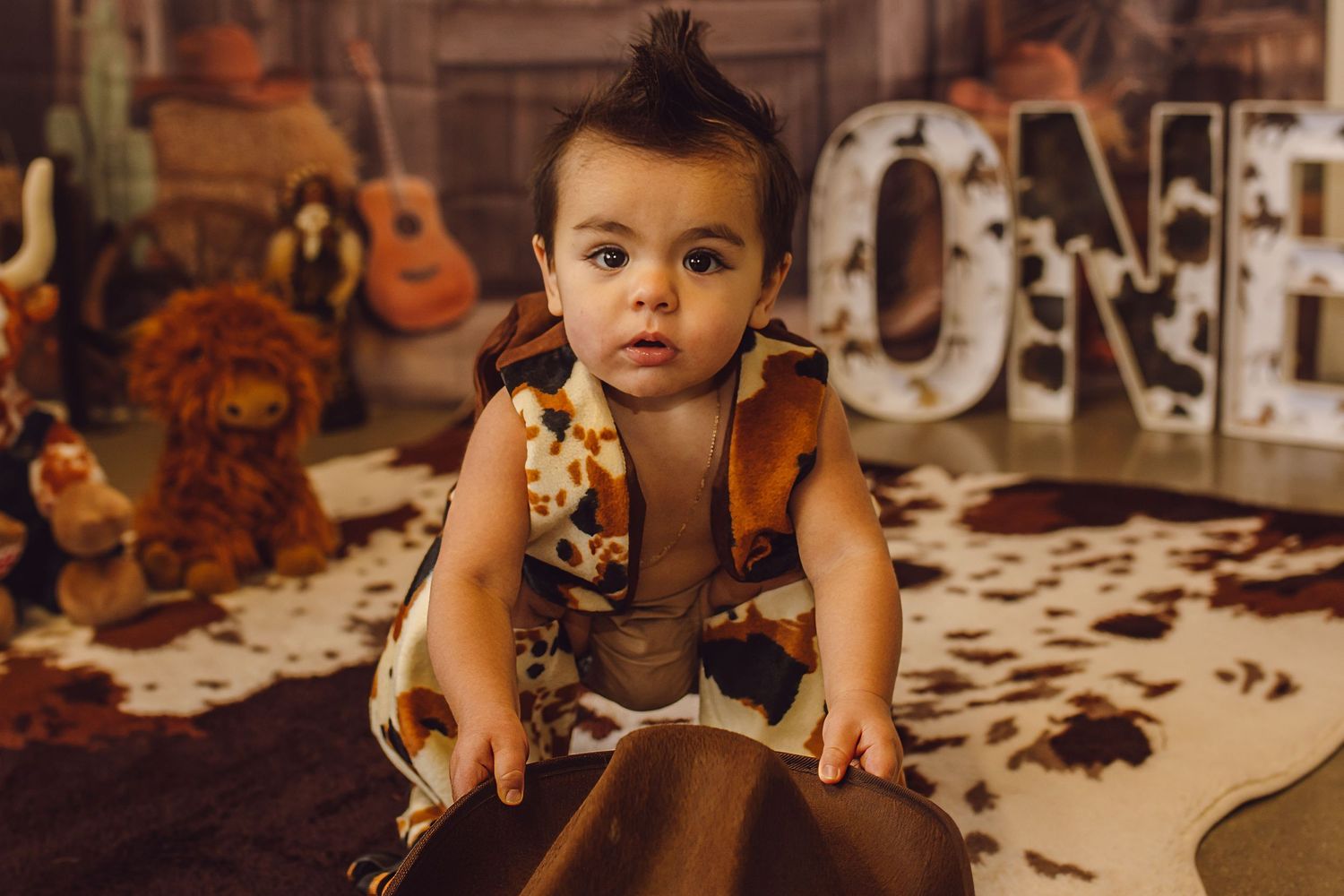 Baby in cow print outfit sits on fur rug near ONE sign and toys at a first birthday photo shoot.