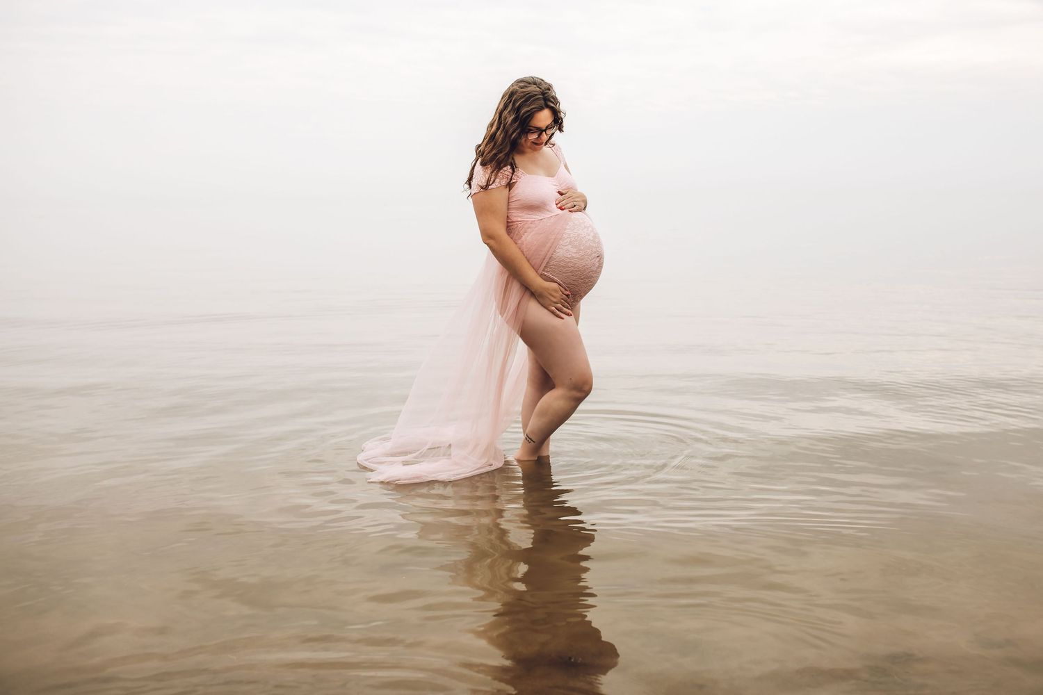 Pregnant woman in sheer pink gown standing in calm water during a serene maternity photo shoot.