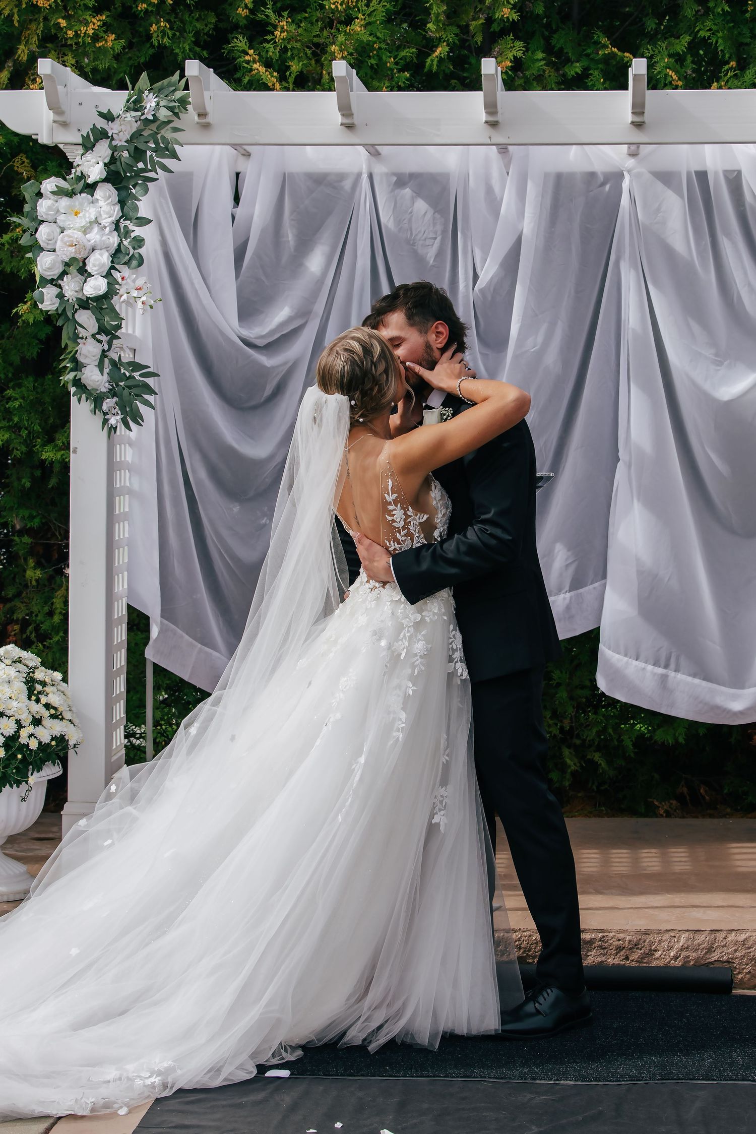 Bride and groom share first kiss at outdoor wedding ceremony beneath white floral arch with draped gray fabric backdrop.