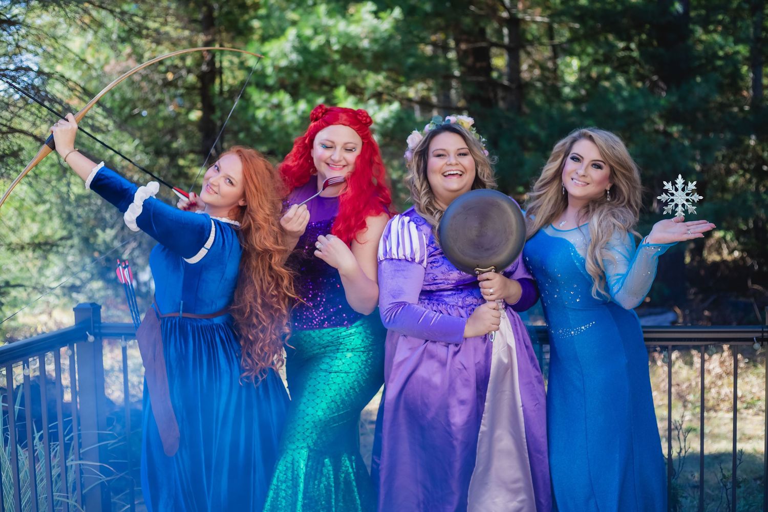 Four women in Disney princess costumes pose outdoors on a deck, dressed as Merida, Ariel, Rapunzel, and Elsa.