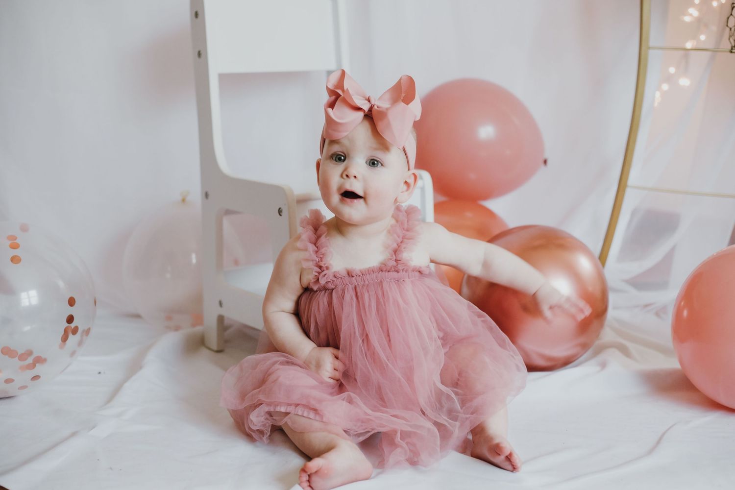 A baby girl in a pink tulle dress and bow headband sits surrounded by pink balloons at her first birthday celebration.