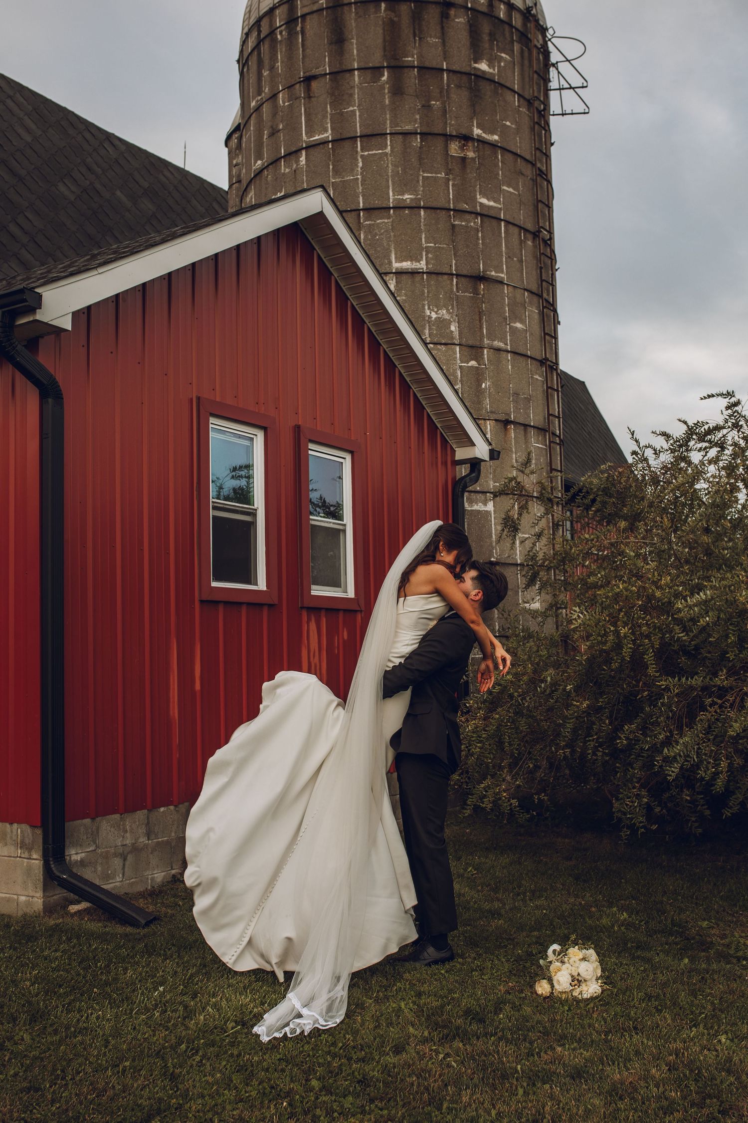 A couple in wedding attire shares a romantic moment against a rustic red barn and stone silo in a countryside setting.