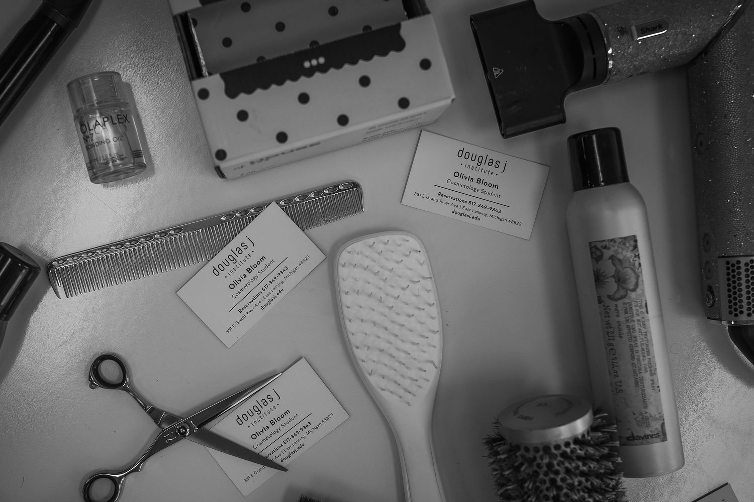 Black and white flat lay of barber tools including scissors, combs, brushes, hair products, and business cards on white surface.